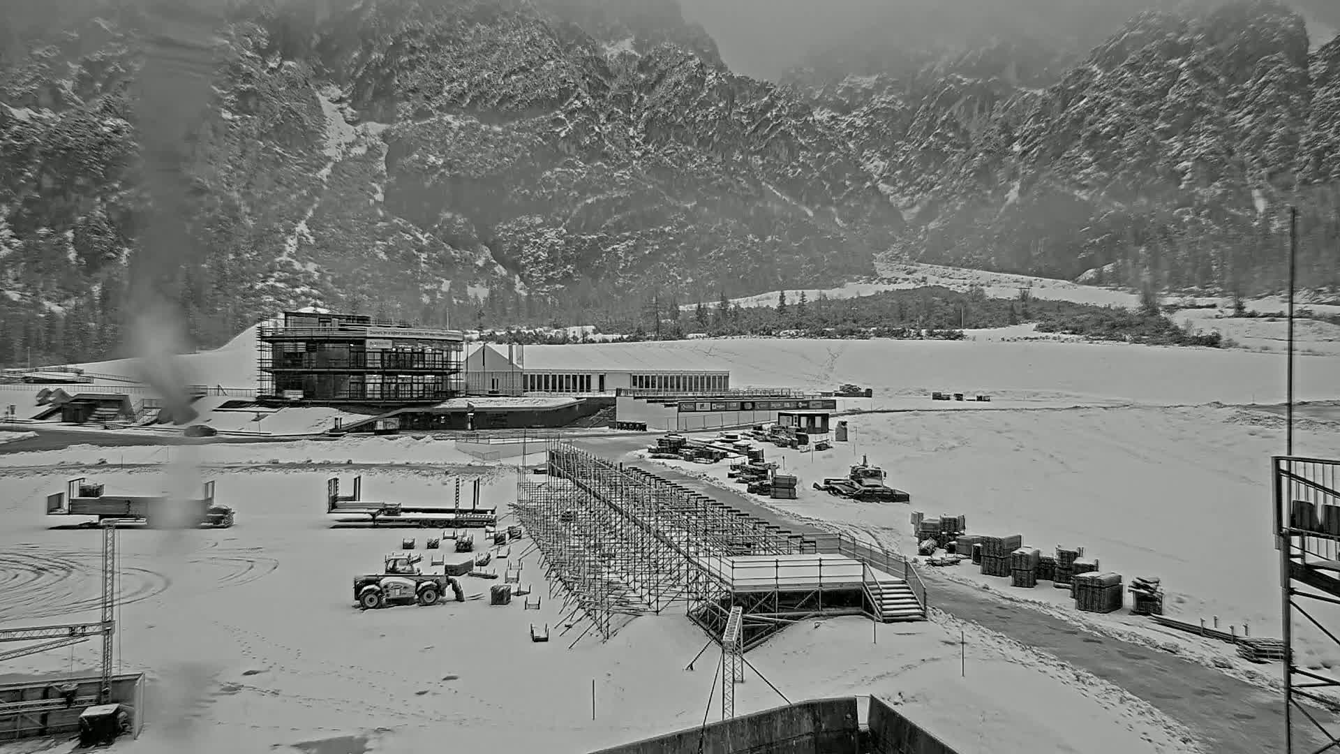 A snowy landscape shows a building complex and construction equipment at the base of a snow-covered mountain range under an overcast sky.