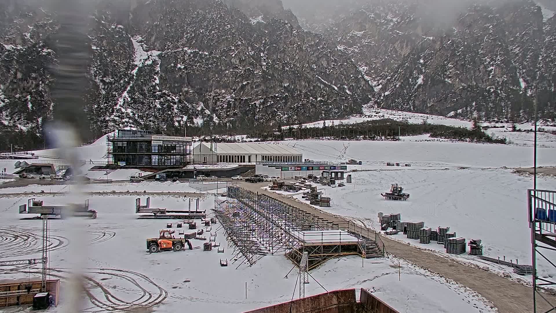 A snowy outdoor scene shows a building complex, construction equipment, and scaffolding in front of a large, snow-covered mountain range under an overcast sky.