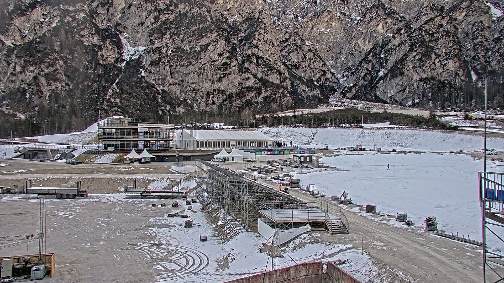 A snowy outdoor scene shows a partially constructed sports facility nestled against a large mountain range, with several trucks and construction materials scattered around the site.