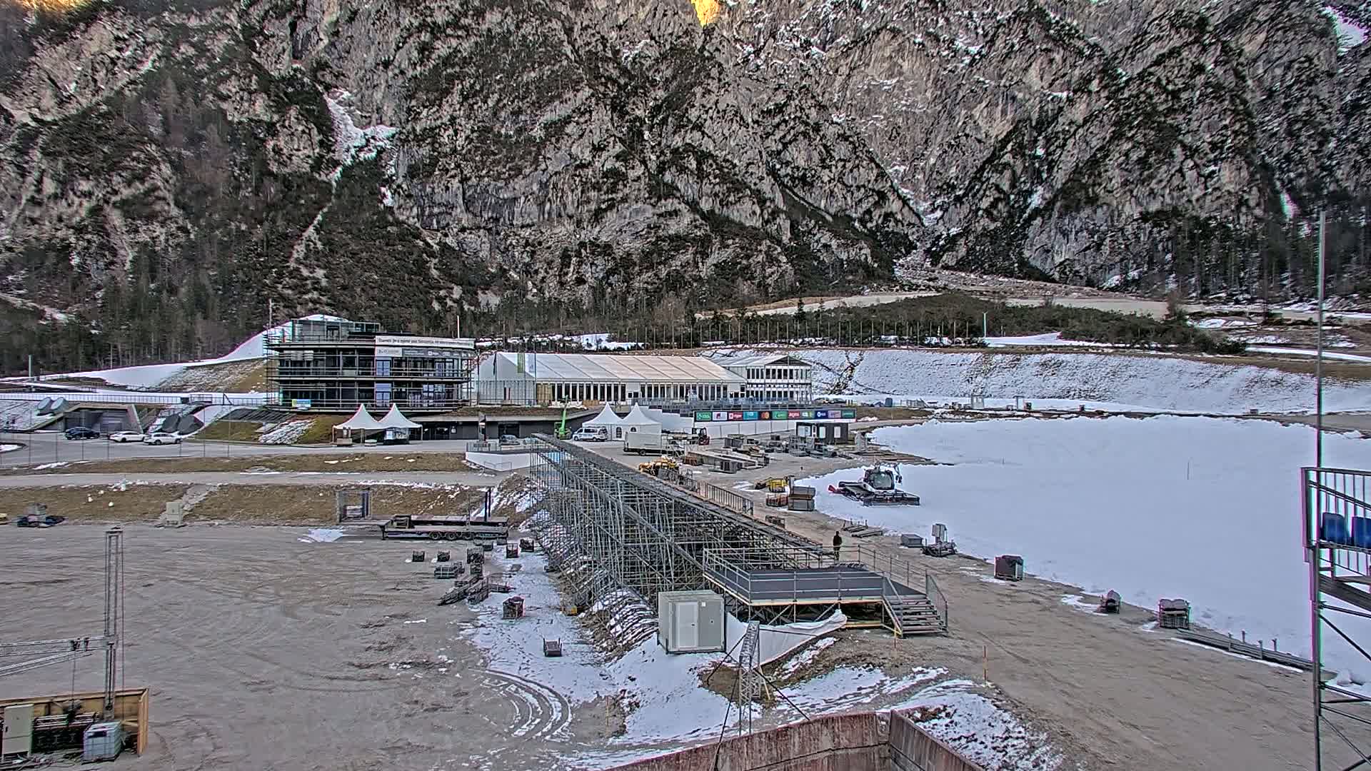 A partially snow-covered outdoor sports complex nestled in a mountain valley, featuring several buildings, temporary structures, and construction equipment.
