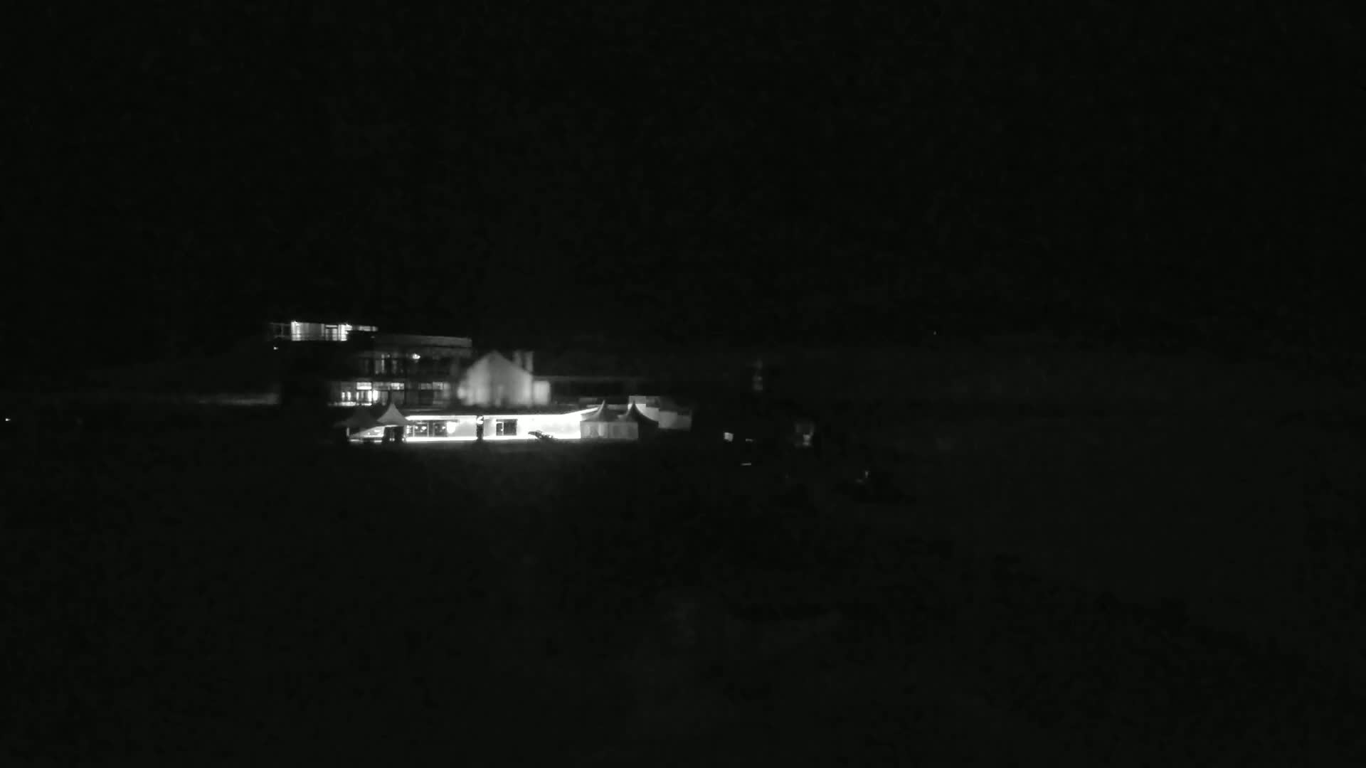 A brightly lit building and some tents are visible at night in a dark, possibly snowy, outdoor setting.