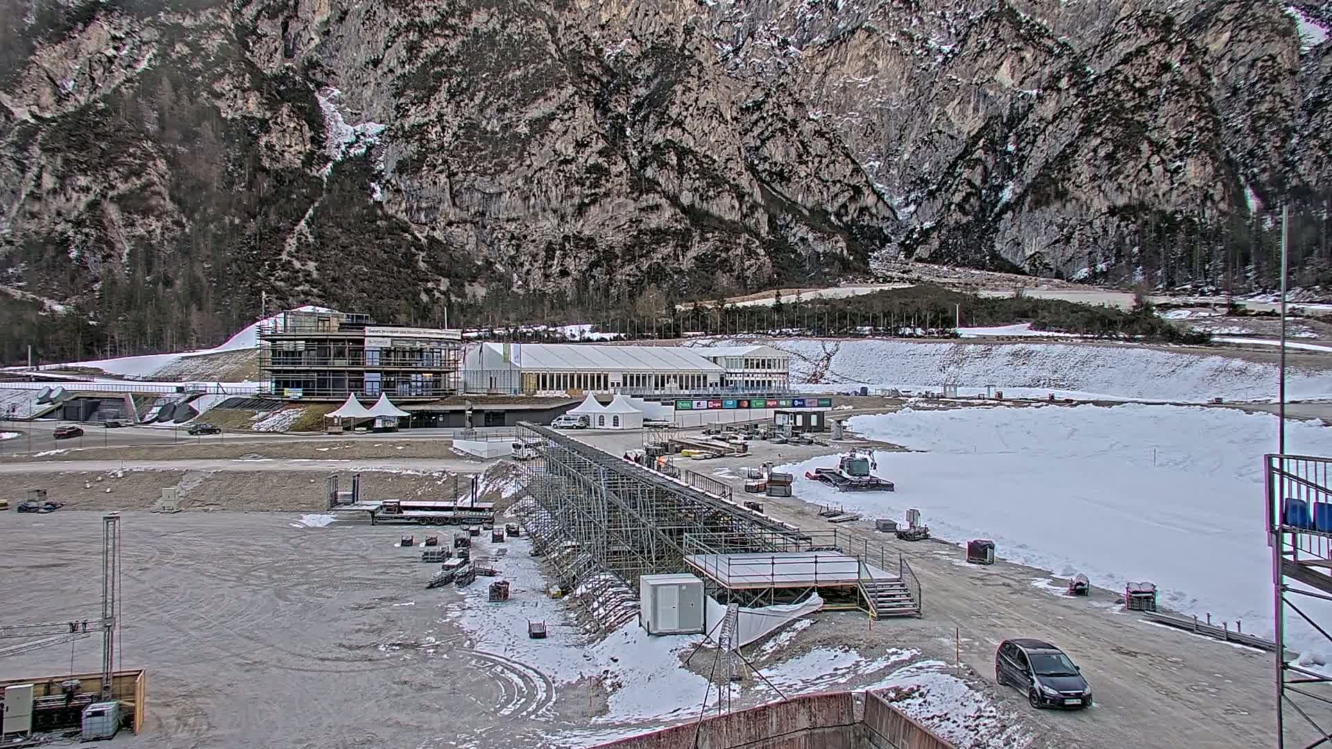 A partially snow-covered outdoor area with several buildings, temporary structures, and construction equipment sits against a backdrop of a large, rocky mountain.