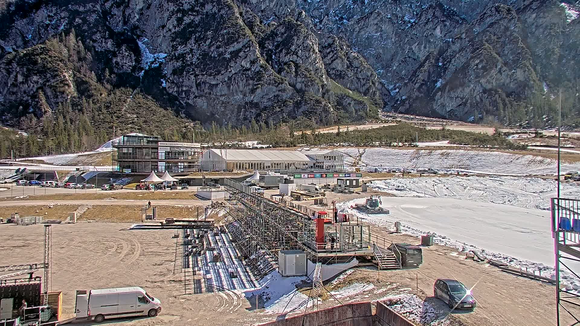 A snowy outdoor sporting venue with buildings, vehicles, and scaffolding is situated against a backdrop of a large mountain range under a sunny sky.