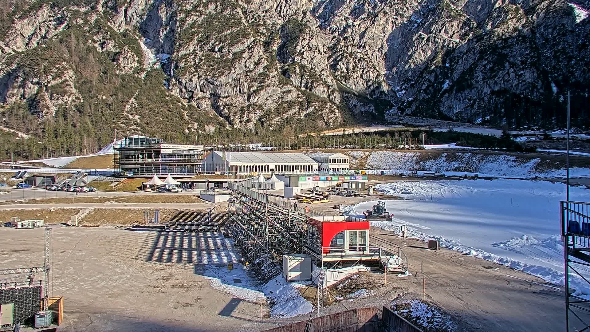 A snowy outdoor sports facility, nestled against a mountainous backdrop under sunny conditions, features several buildings, including a large structure and several temporary tents, along with various equipment and staging areas.