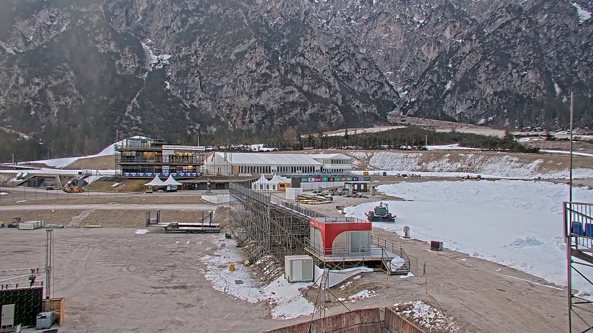 A partially snow-covered outdoor area with buildings under construction,  a large temporary structure, and snow-grooming equipment, set against a backdrop of a mountainous landscape.