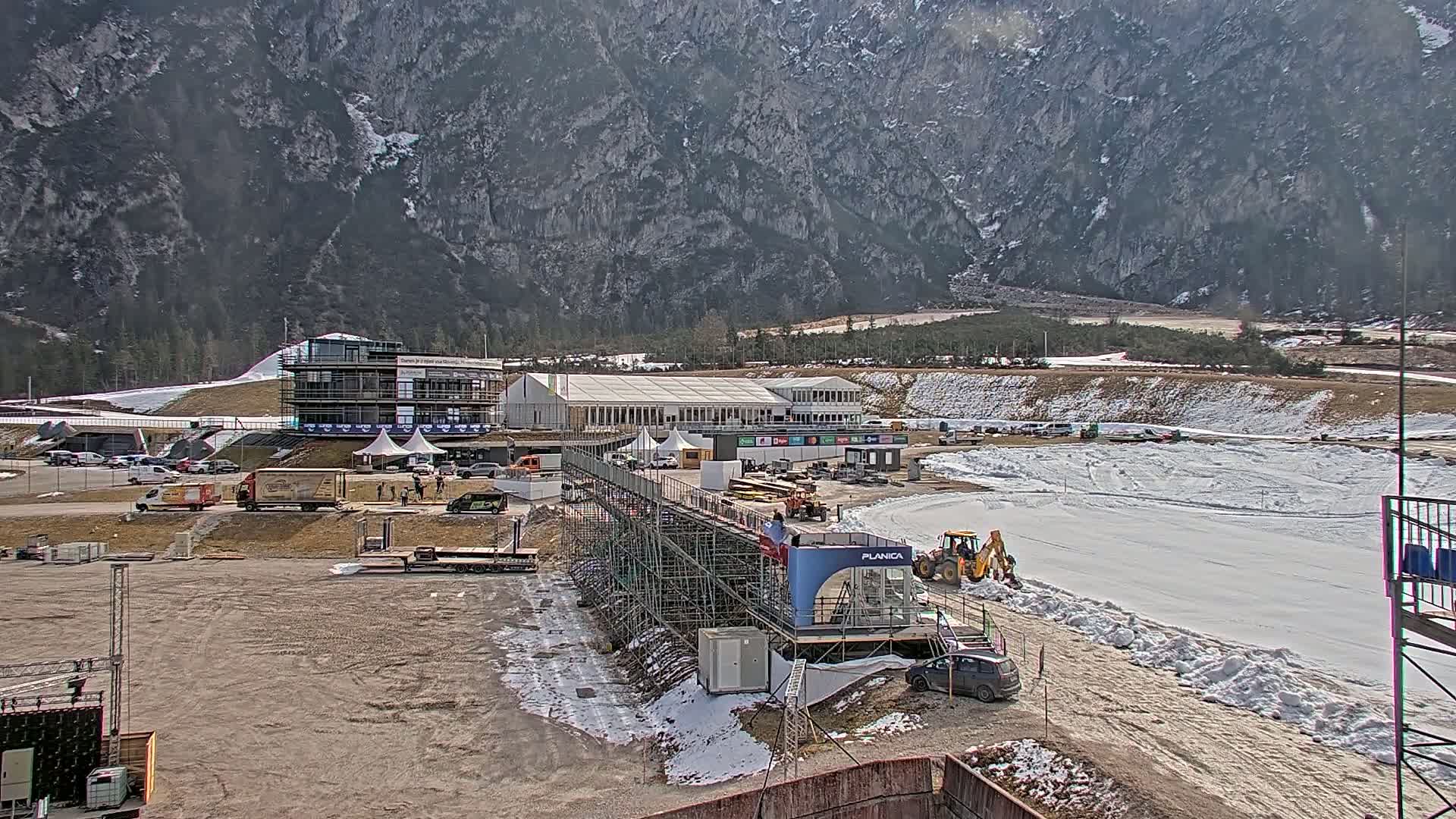 A ski jumping facility under construction is situated in a valley, backed by a large mountain range on a partly sunny day with patches of snow on the ground.