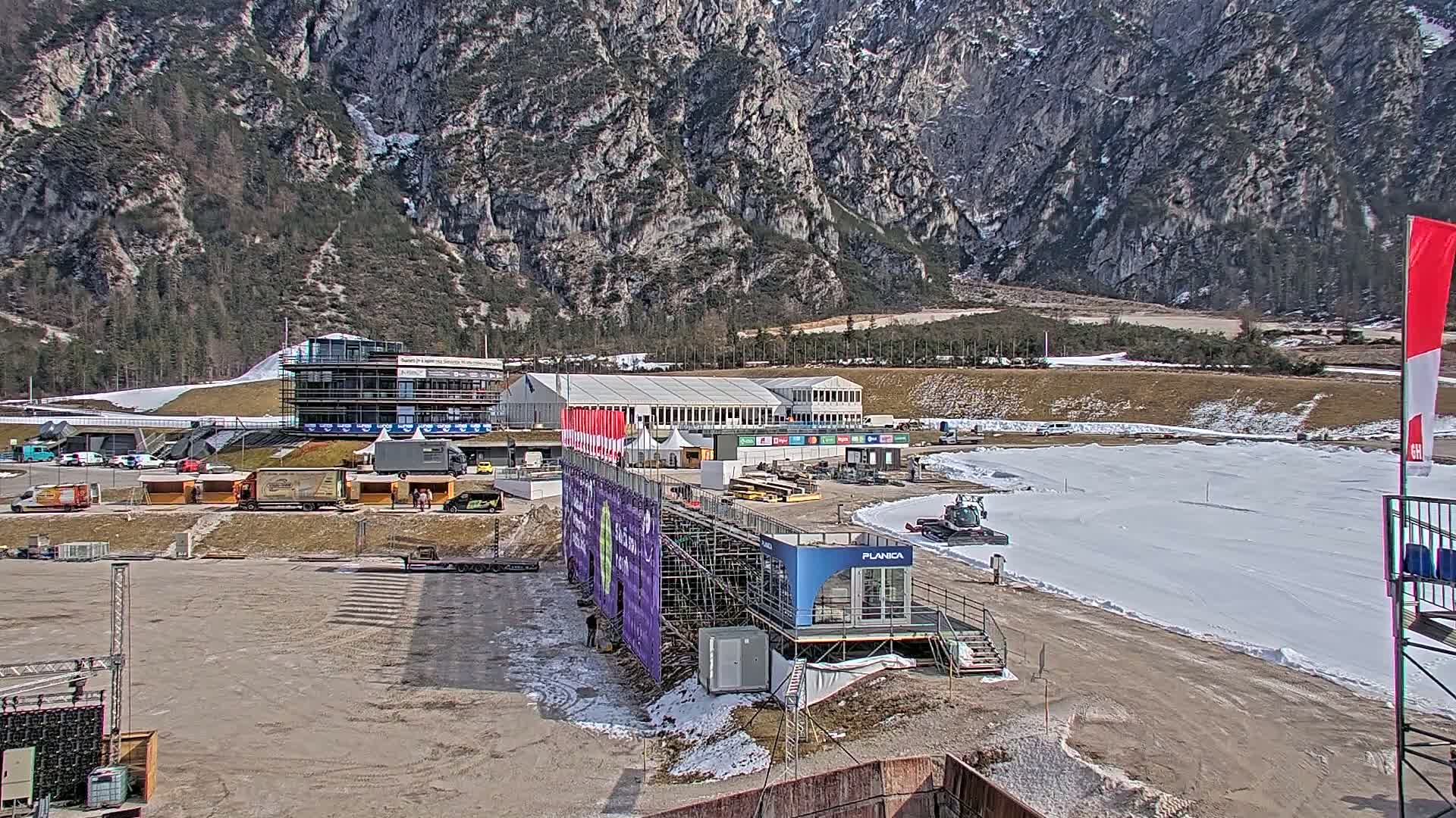 A snow-covered ski jump arena is situated in a valley below a large, rocky mountain range on a mostly sunny day.