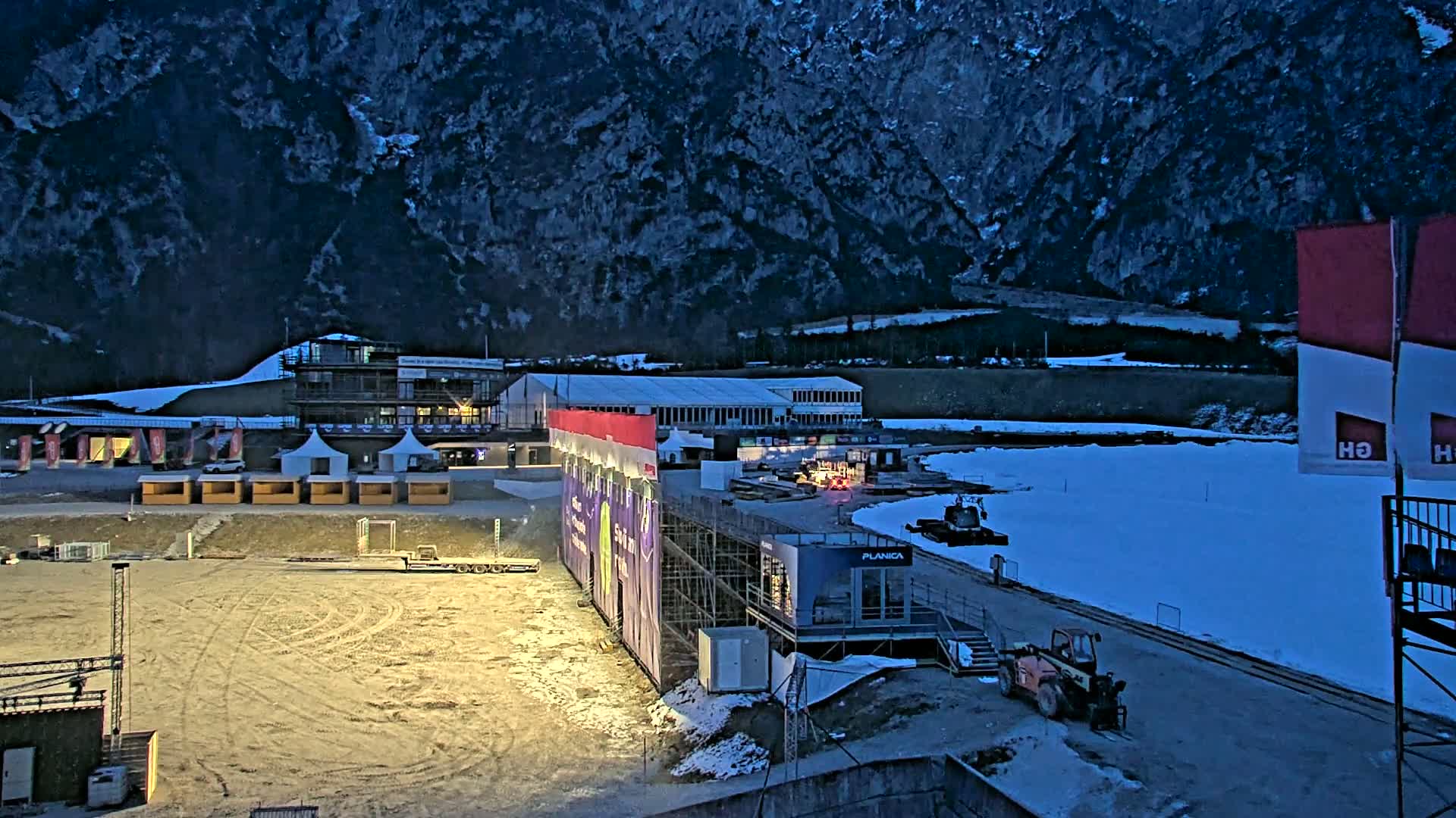 A snow-covered outdoor arena at dusk, nestled against a dark mountainside, features various temporary structures, including tents and scaffolding, with some machinery present.