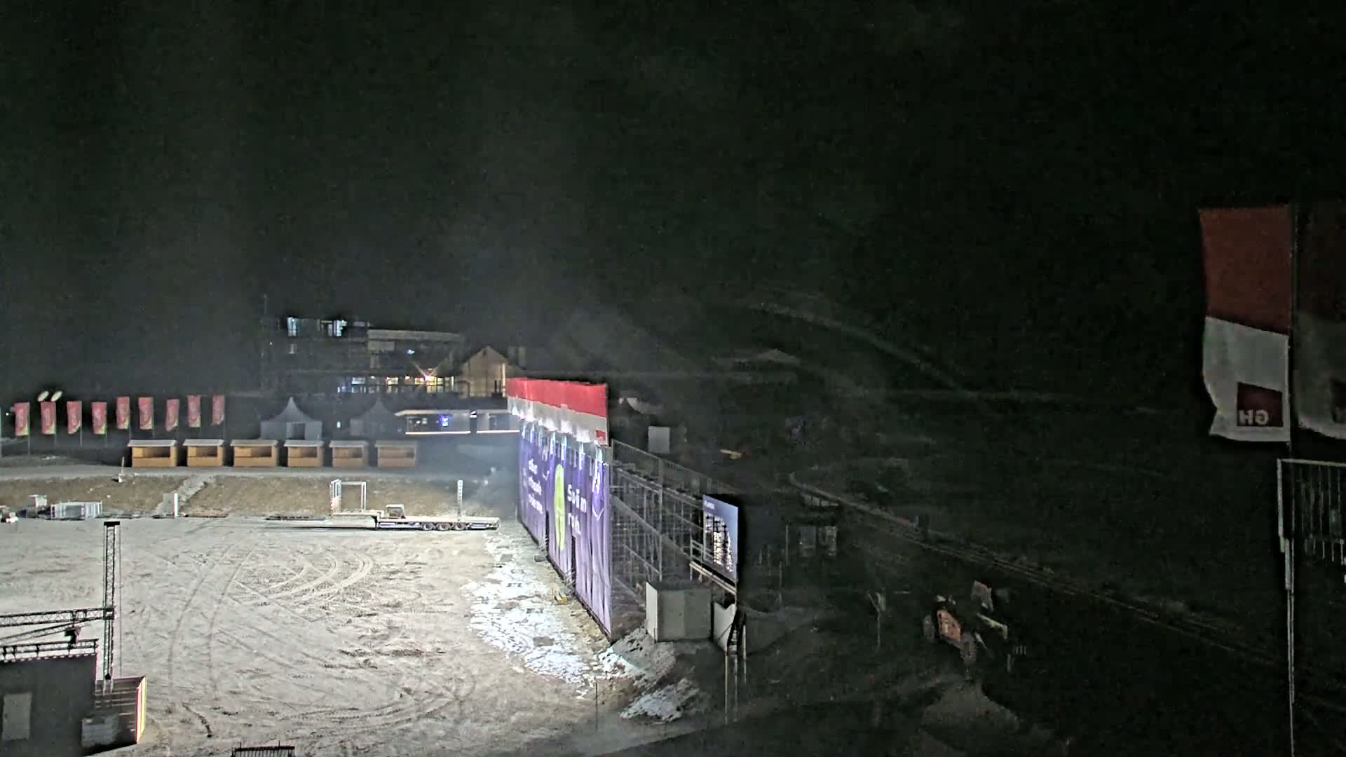 A snow-covered, nighttime outdoor area features a large purple wall with a scoreboard, several small structures, and a building in the background under a dark, possibly snowy sky.