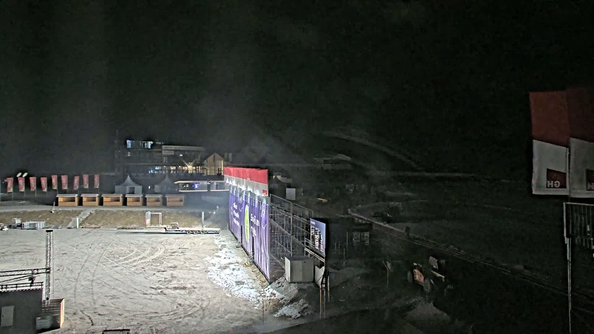 A snow-covered nighttime outdoor area features a large structure with a purple banner, small wooden huts, and a building in the background under dark, hazy conditions.