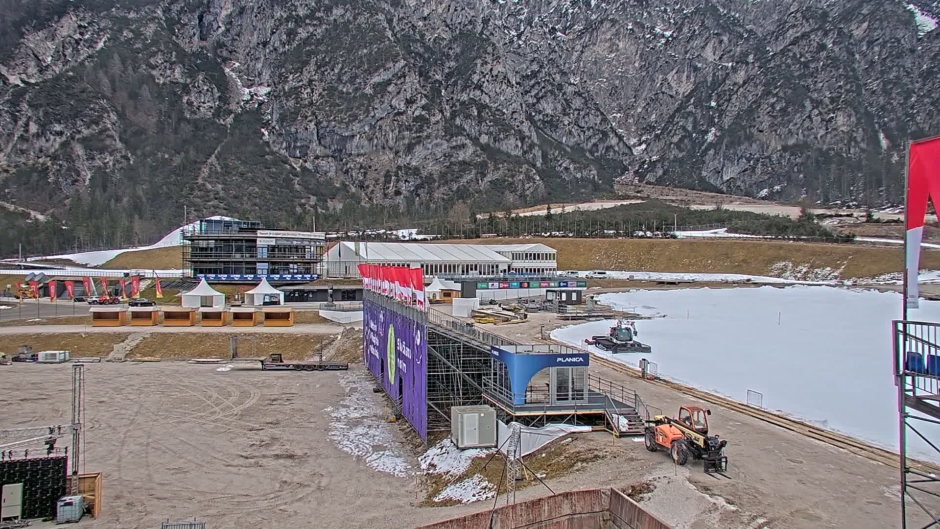 An outdoor sports complex is partially covered in snow, situated against a backdrop of a large mountain range under a cloudy sky.