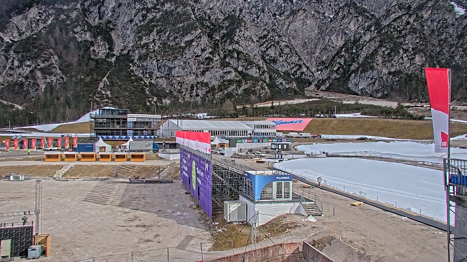 A partially snow-covered outdoor sporting venue is situated at the foot of a large mountain, under a clear sky.