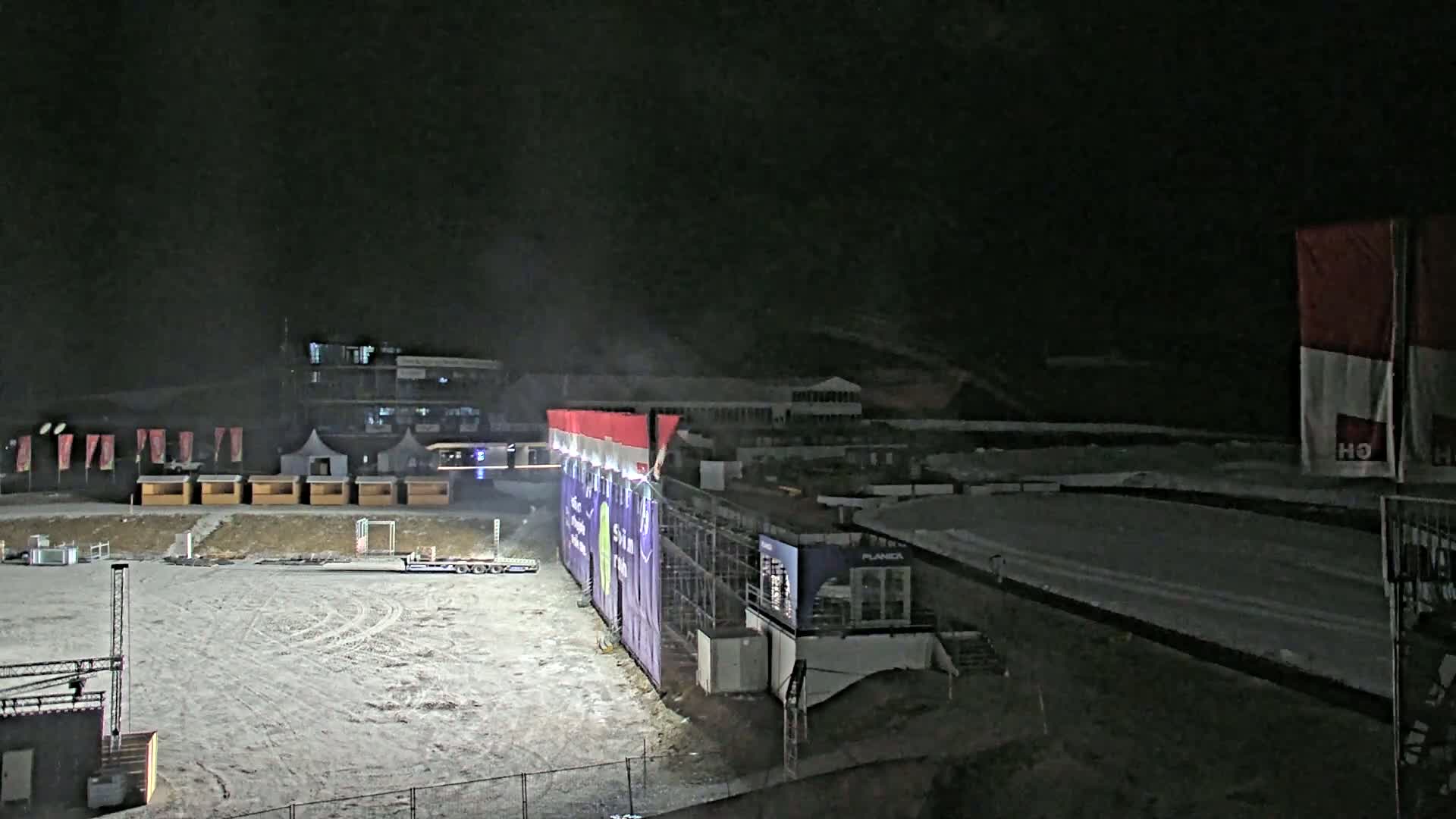 A snow-covered outdoor arena at night is illuminated, featuring a large structure with a screen, small vendor booths, and equipment.