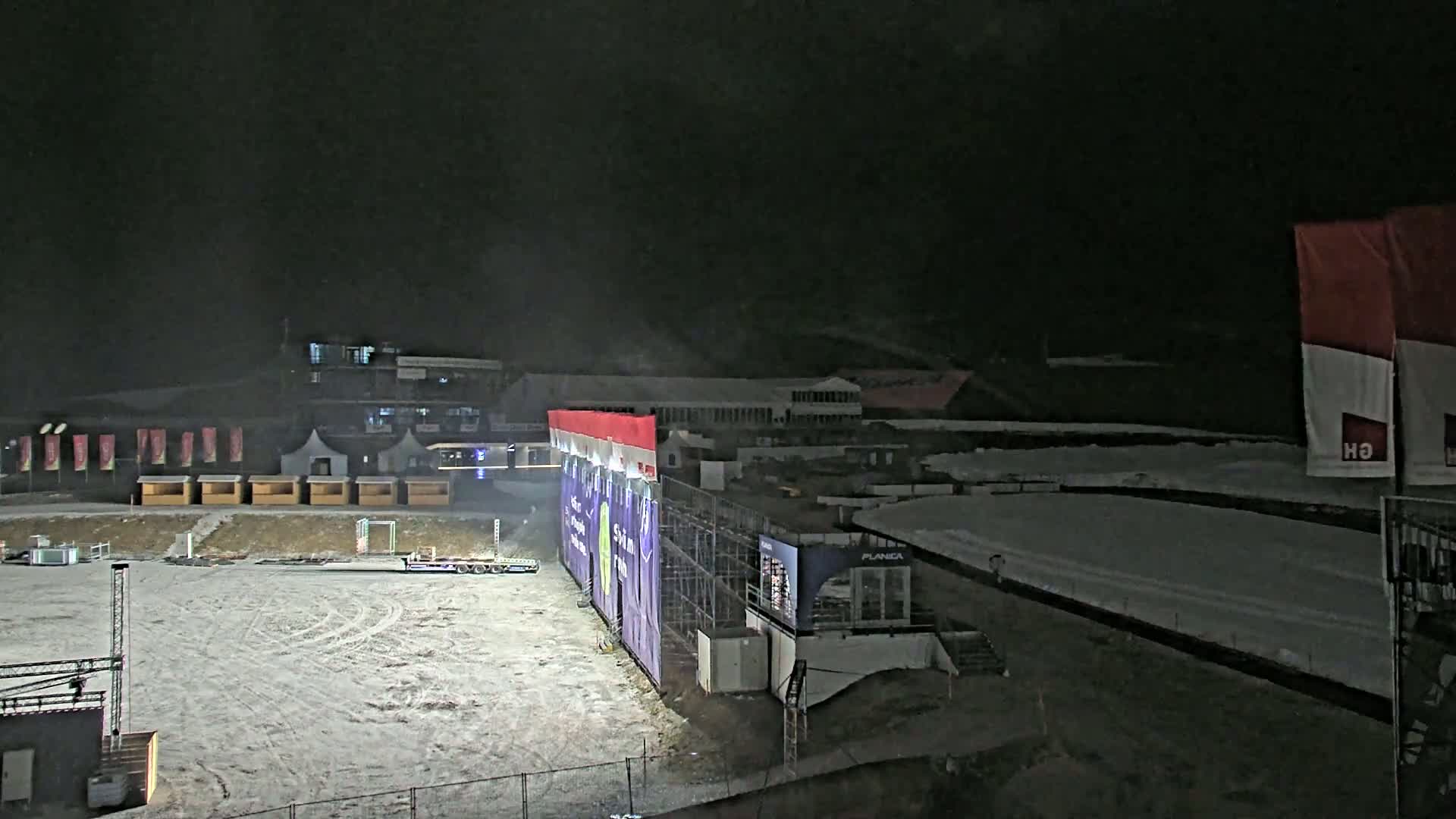 A snow-covered outdoor area at night features a large structure with purple and red signage, smaller buildings in the background, and several small structures and a flatbed trailer in the foreground.