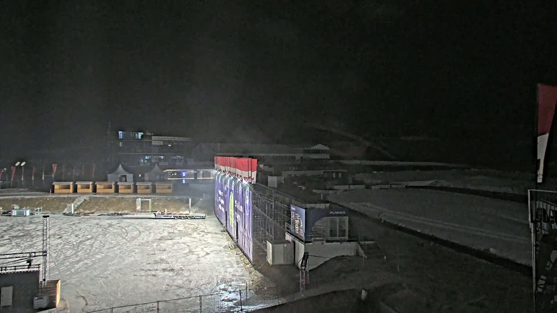 A snow-covered nighttime outdoor arena with temporary structures, a large billboard, and a few vehicles.