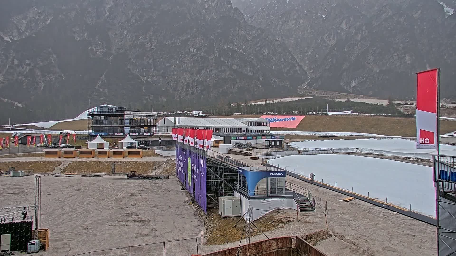 An overcast day reveals a ski jumping venue with snow-covered ramps, several temporary structures, and large banners, all set against a backdrop of mountainous terrain.