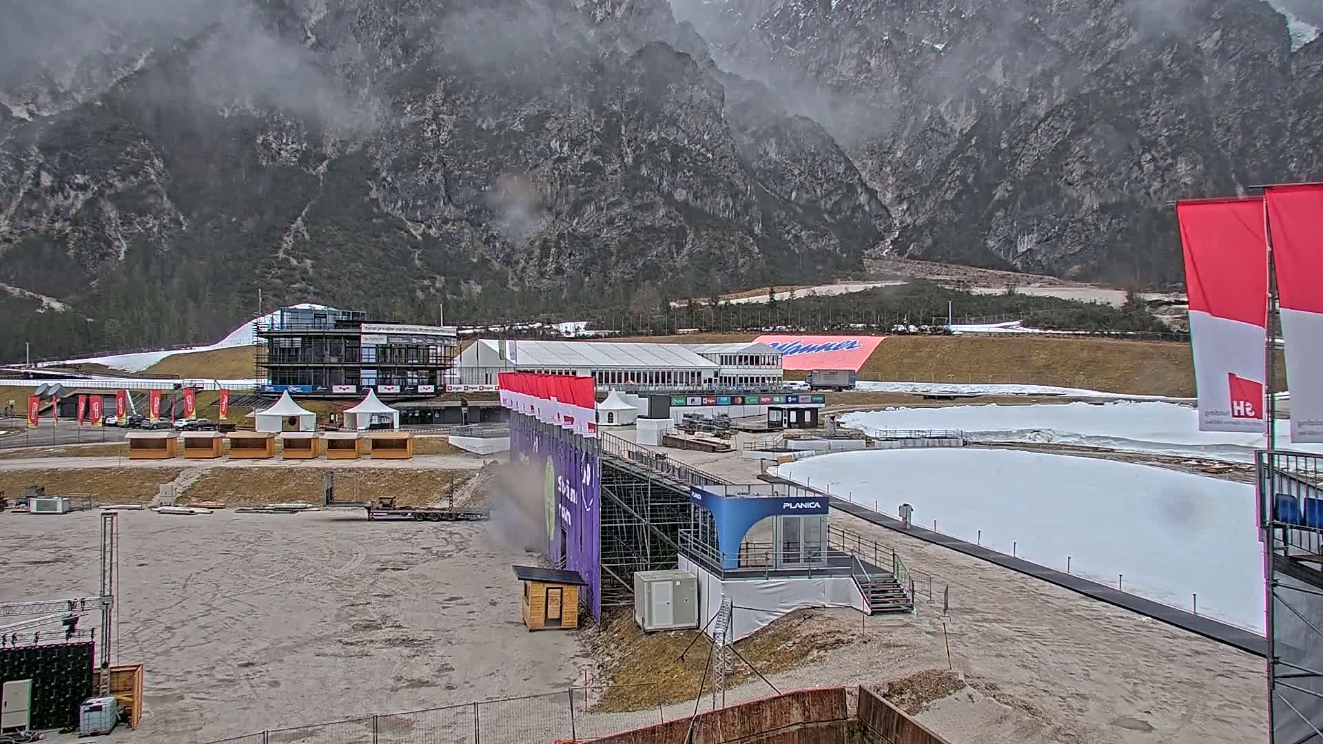 A snowy ski jumping facility sits in a valley overlooked by a mountain range partially obscured by fog.