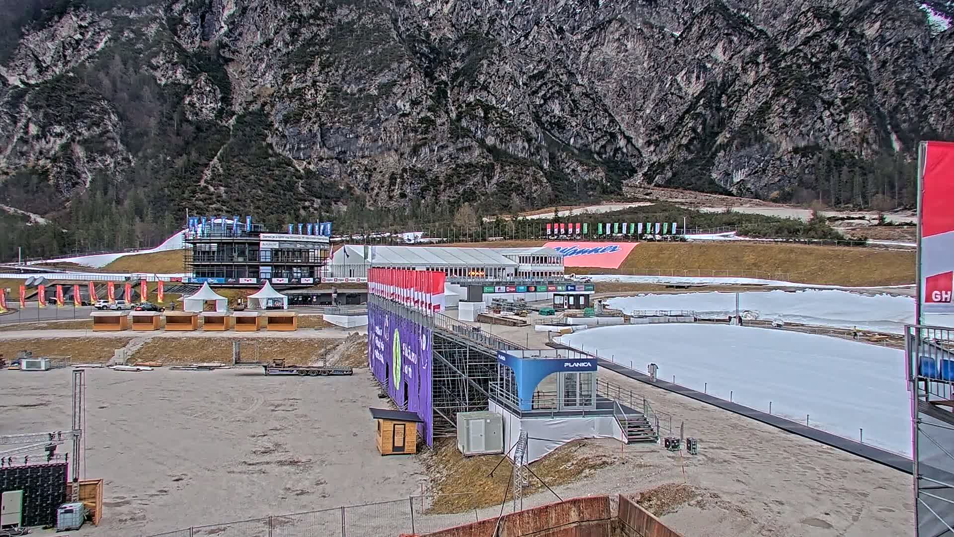 An outdoor ski jumping facility is partially constructed, situated against a backdrop of rocky mountains under overcast skies.