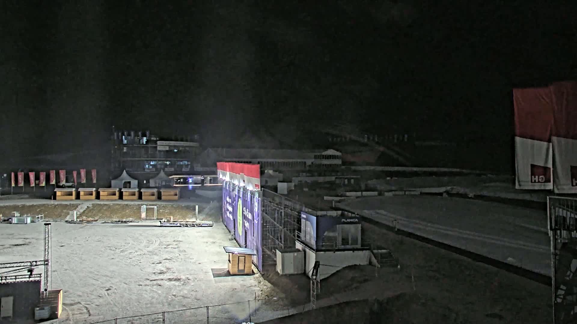 A snow-covered outdoor area at night, featuring several small structures, a large purple banner, and a building in the background.