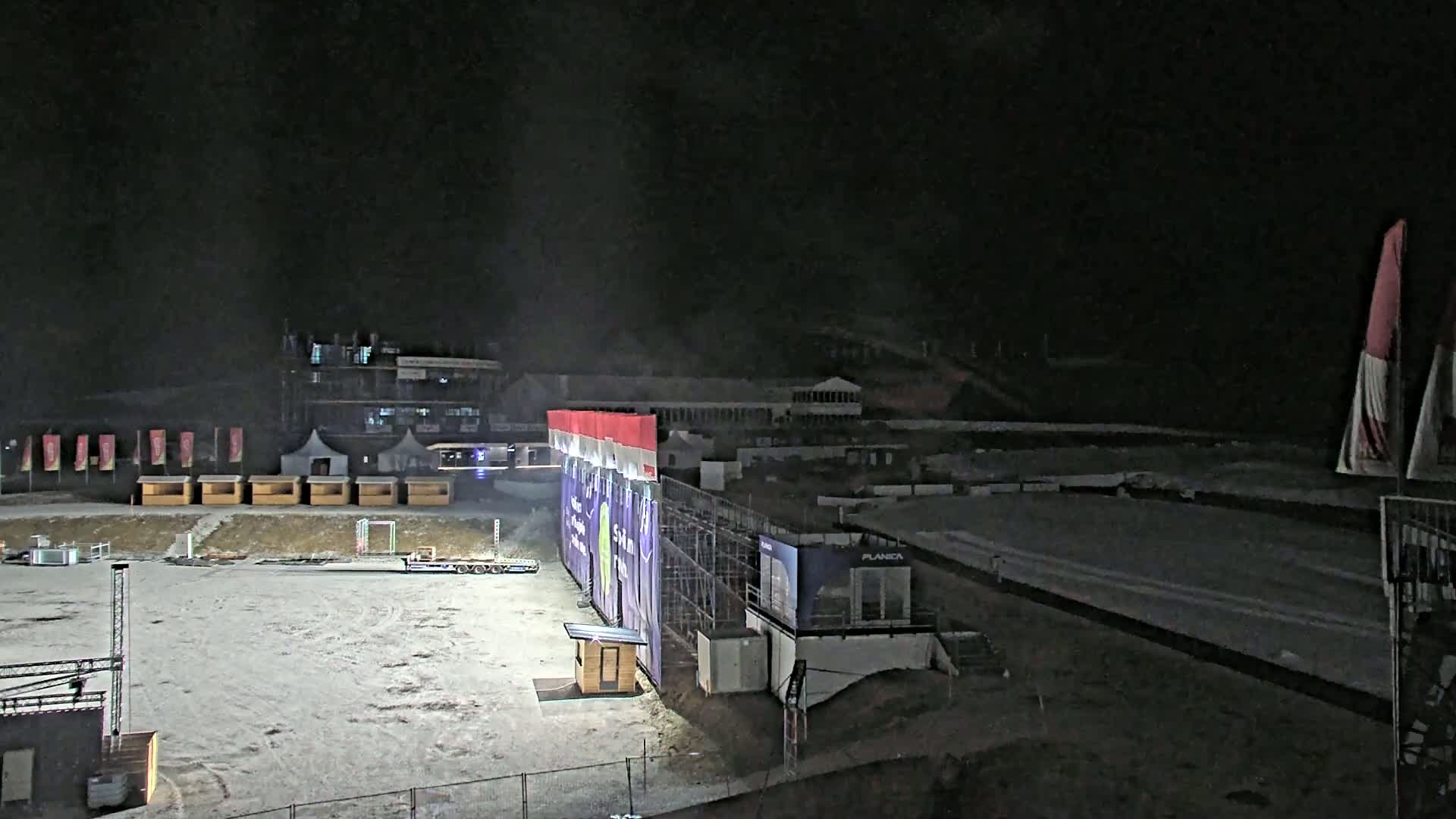 A snow-covered outdoor area at night with several small buildings, a large structure with banners, and a flat, cleared space.