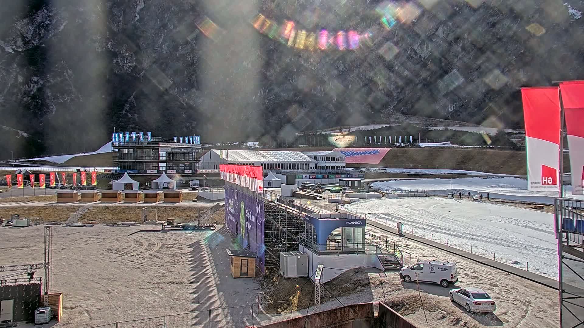A sunny outdoor view of a ski jumping facility with various buildings, temporary structures, and vehicles, partially covered in snow.