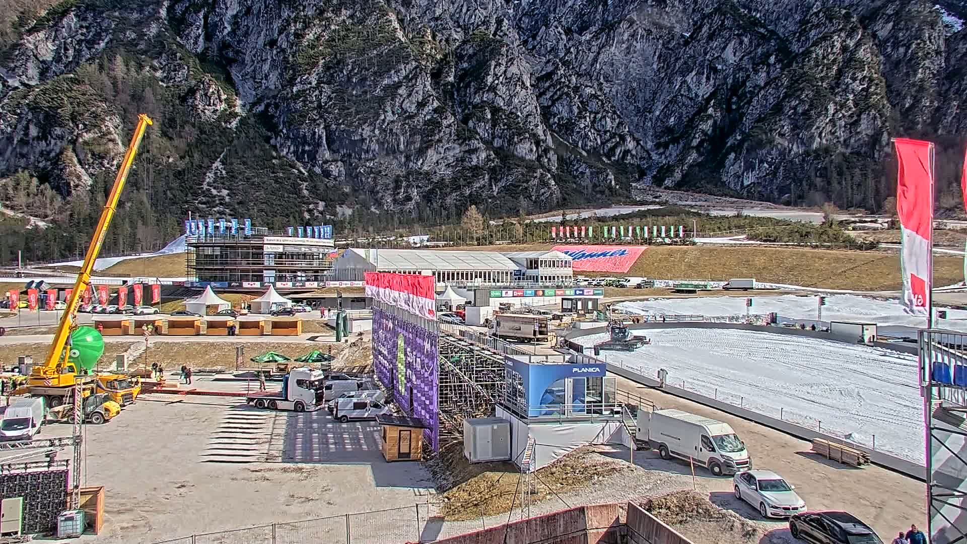 A ski jumping facility with various vehicles, temporary structures, and construction equipment is situated against a mountainous backdrop on a partly sunny day.