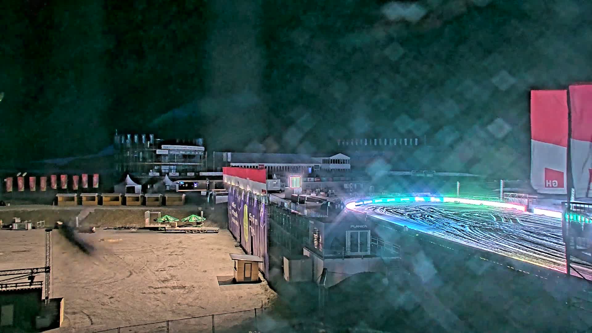 A snow-covered nighttime ski jump track with colorful lights is visible, with surrounding buildings and structures under a hazy, dark sky.