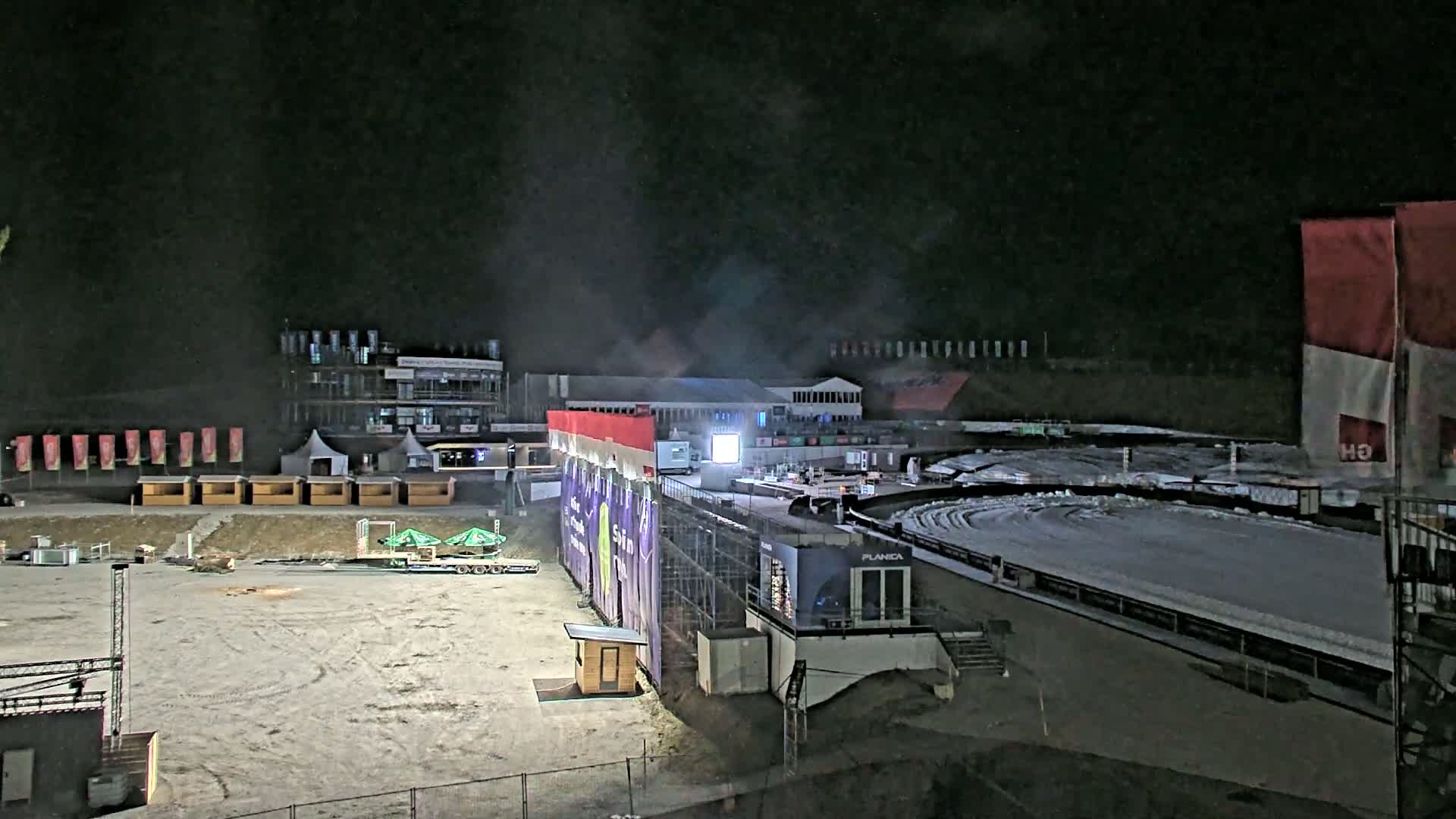 A nighttime view of a snow-covered outdoor sports arena with various structures and equipment, including temporary buildings and a large screen.