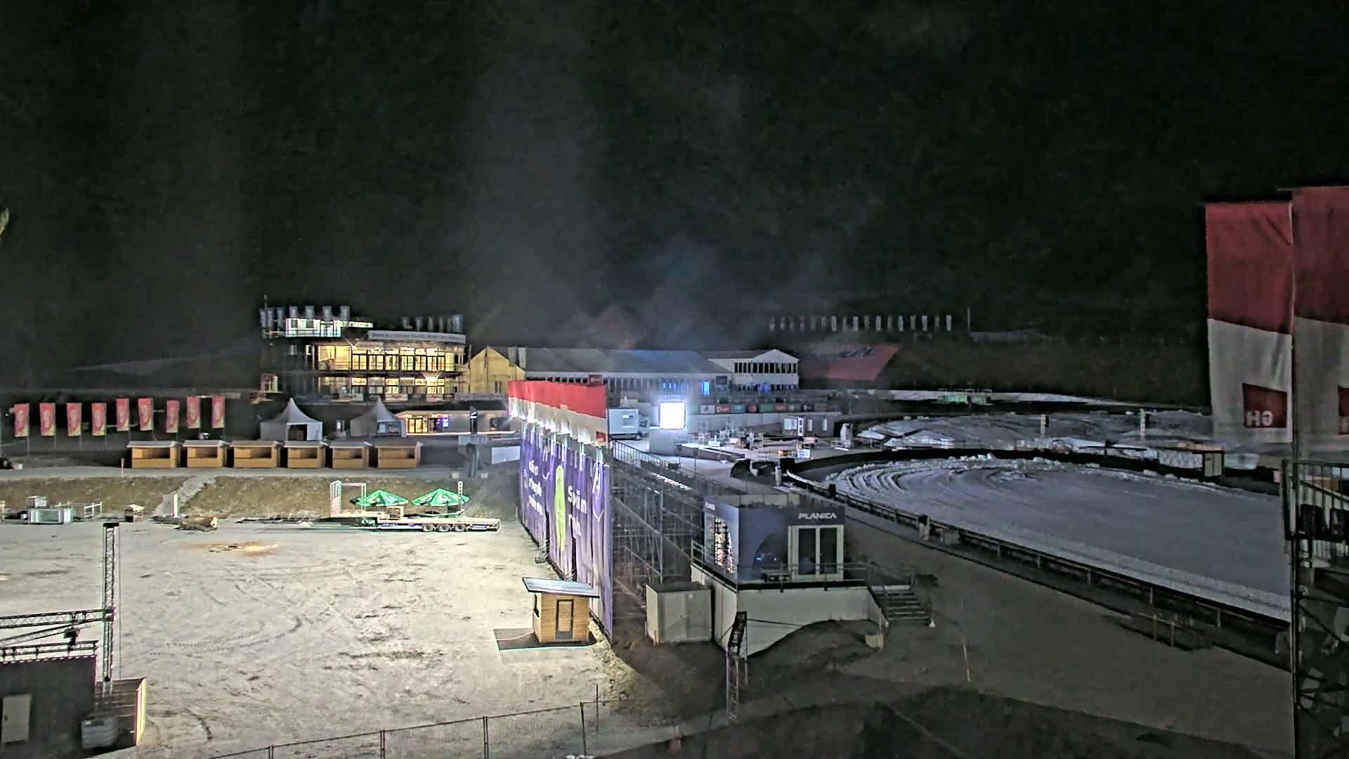 A nighttime view of a snow-covered outdoor arena with buildings and temporary structures surrounding a lit track.