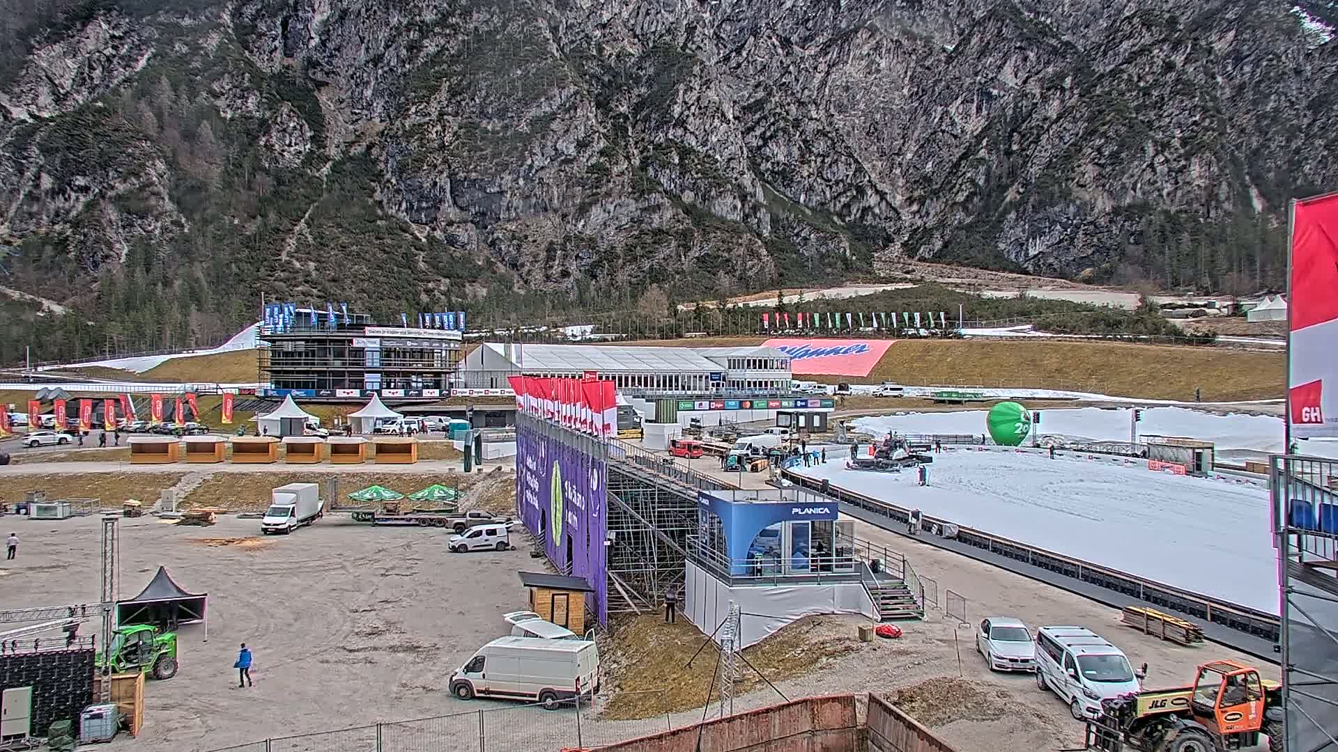 A ski jump stadium with snow-covered areas, numerous vehicles, temporary structures, and a large mountain backdrop in overcast conditions.