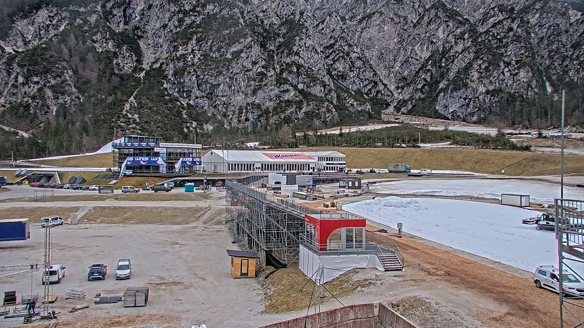 A snowy outdoor sports facility, nestled against a large mountain, shows a building, vehicles, and scaffolding under a cloudy sky.