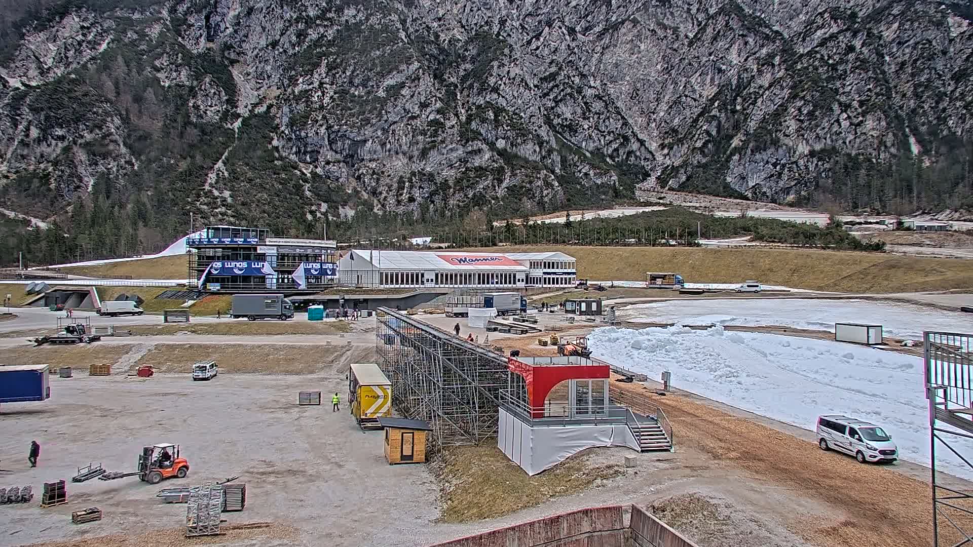 A snowy outdoor track sits in front of a large building against a mountainous backdrop under a cloudy sky.