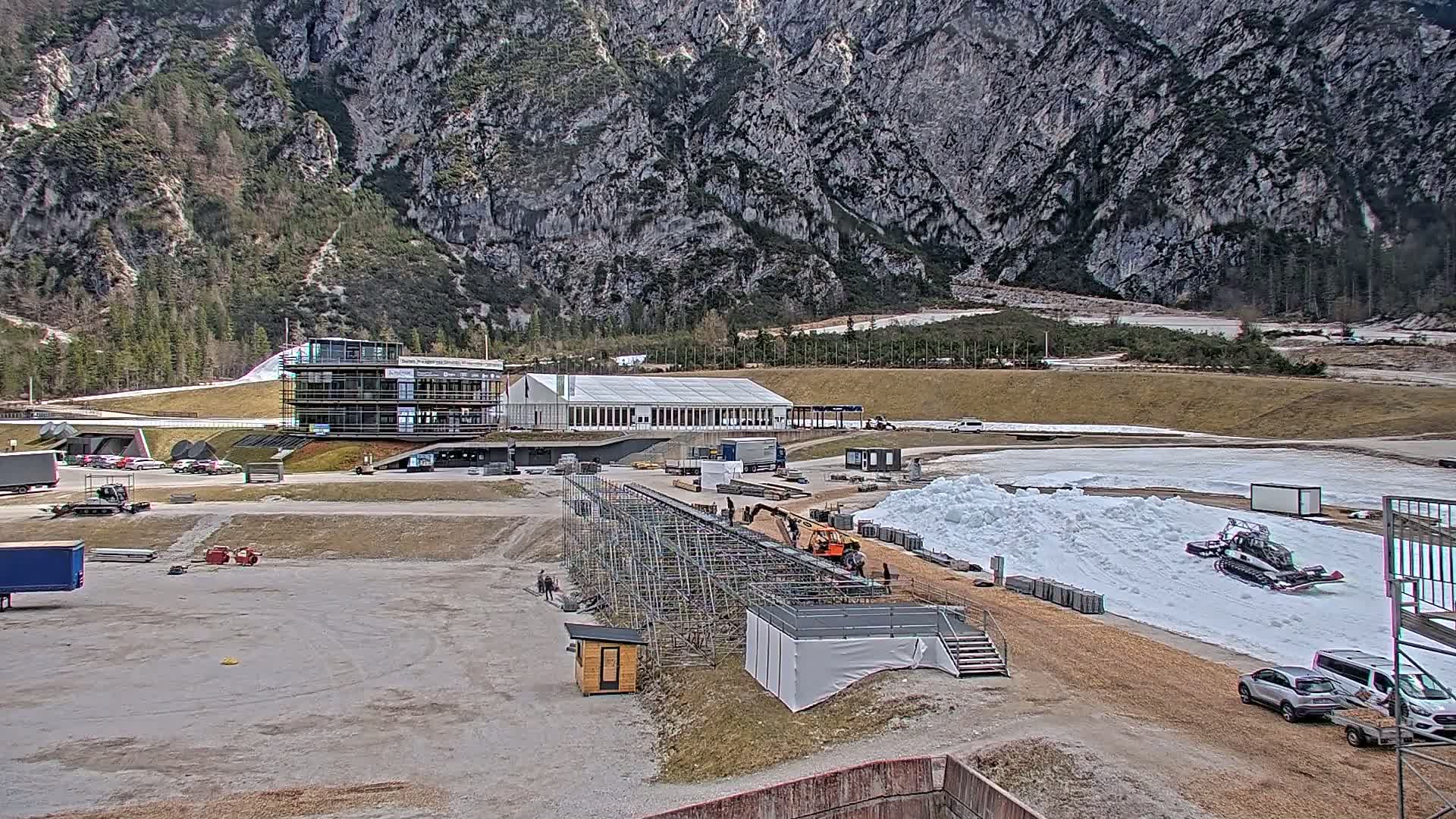 A large, grey mountain looms behind a partially constructed outdoor sports facility with a snow-grooming machine working on a snow track, under a mostly cloudy sky.