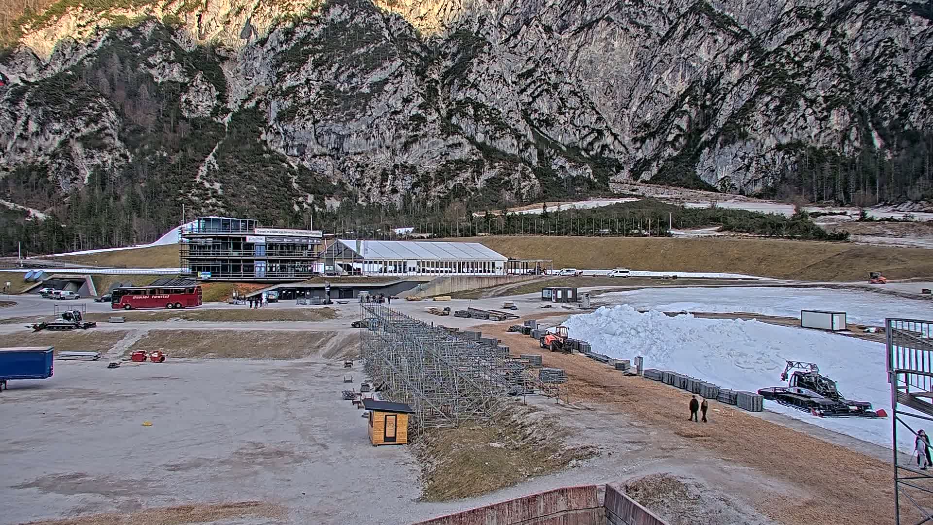 A snow-grooming machine is working on a snow track in a mountain valley, near a building complex and several vehicles.