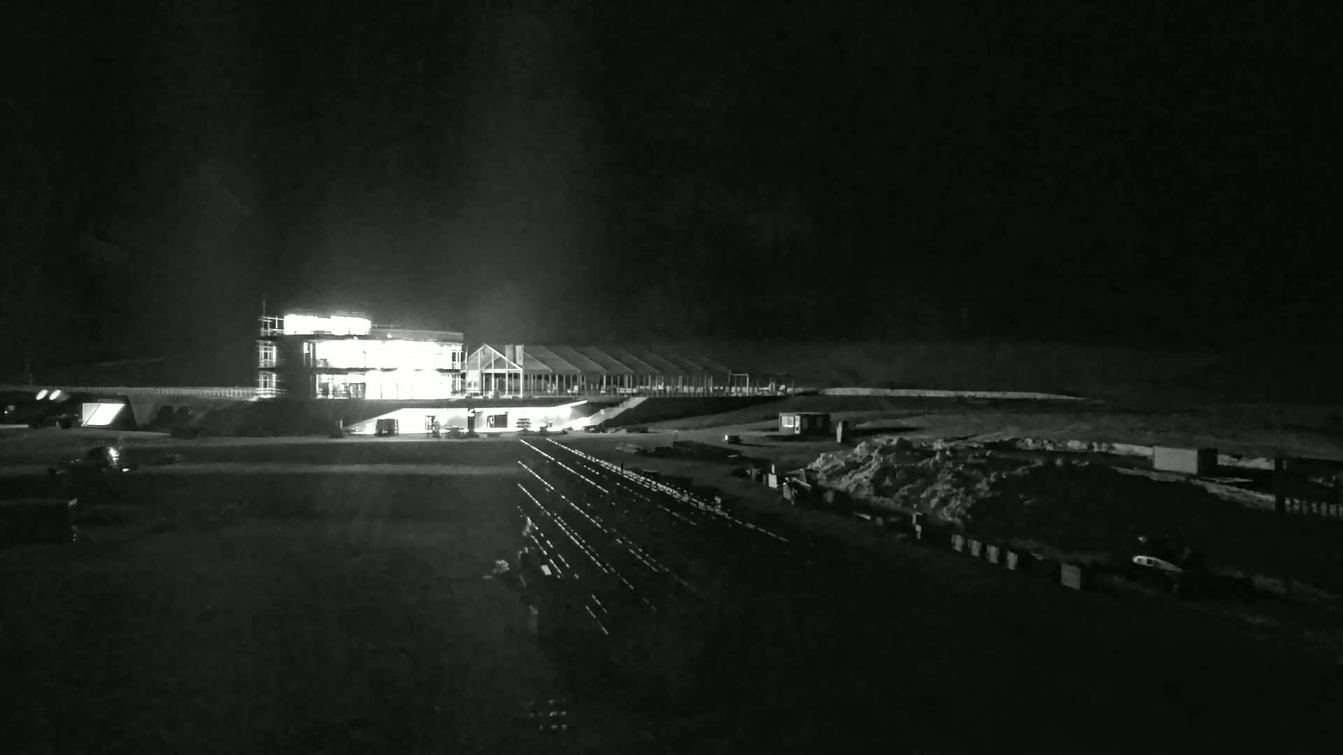 At night, a brightly lit building and a row of smaller structures are visible, with rows of what appear to be bleachers or seating in the foreground, and some snow or piles of material in the lower right.