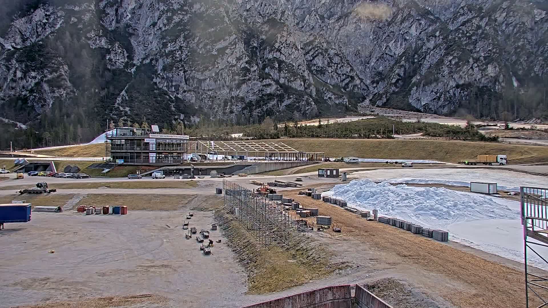 A partially constructed building complex sits in a valley, with piles of snow and construction materials scattered around, against a backdrop of a large, rocky mountain under a partly cloudy sky.