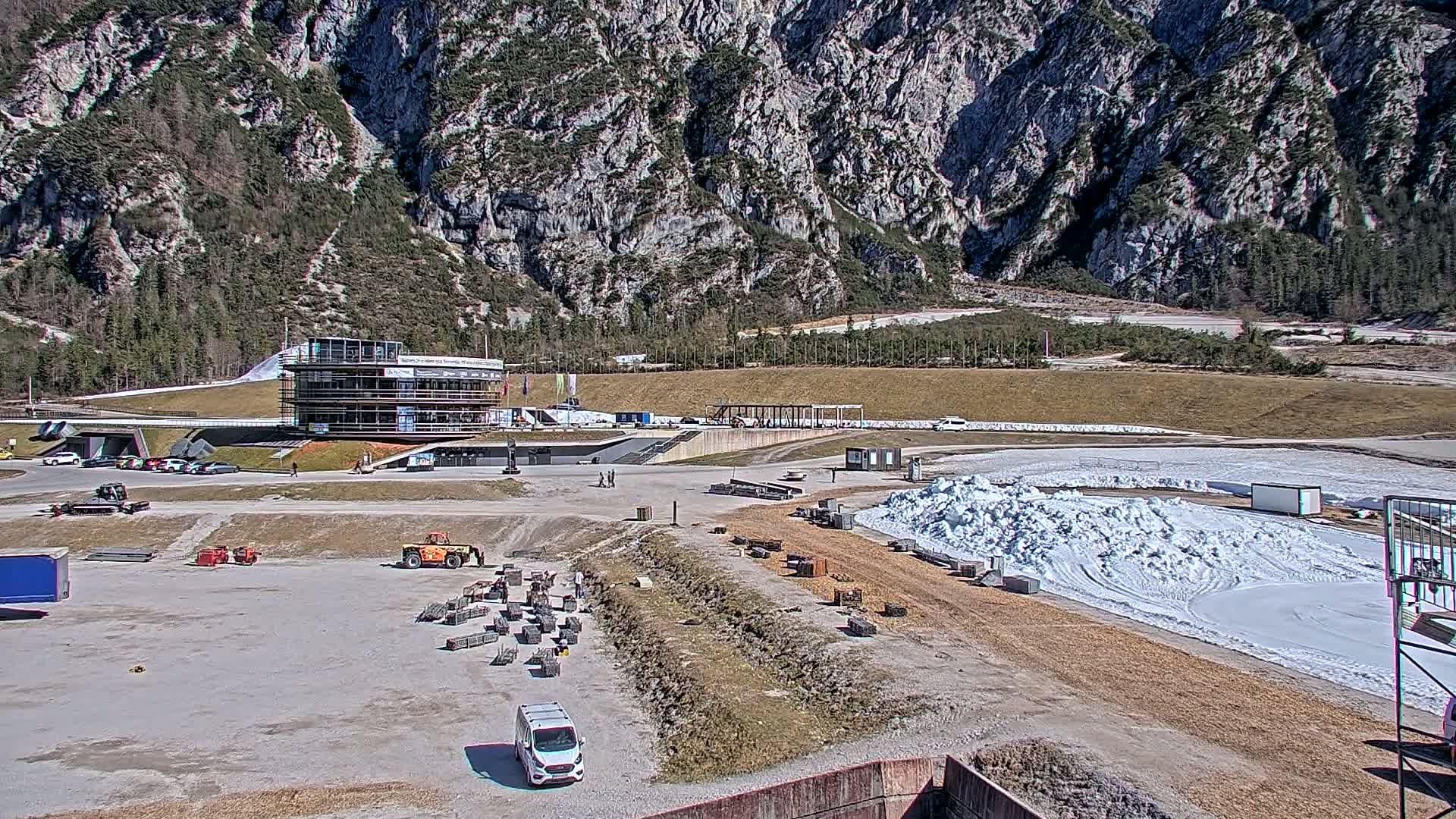 A sunny day reveals a snow-covered track nestled in a valley between imposing mountains, with various vehicles and construction materials scattered around a multi-story building.