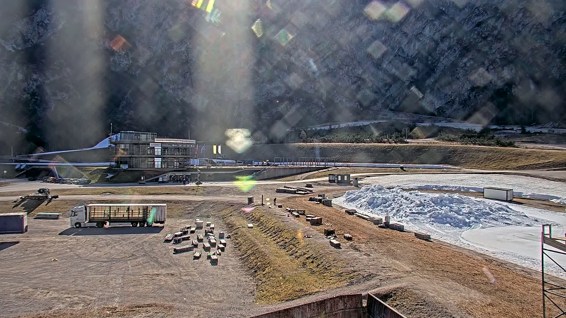 A sunny day reveals a bobsled track nestled in a mountainous region, with a large building and various equipment scattered around the course.