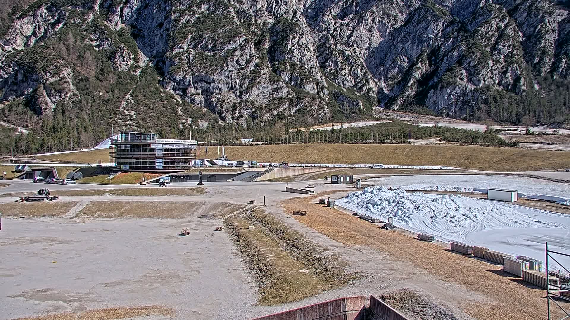 A multi-story building sits near a snowy track against a backdrop of a large, rocky mountain under a sunny sky.