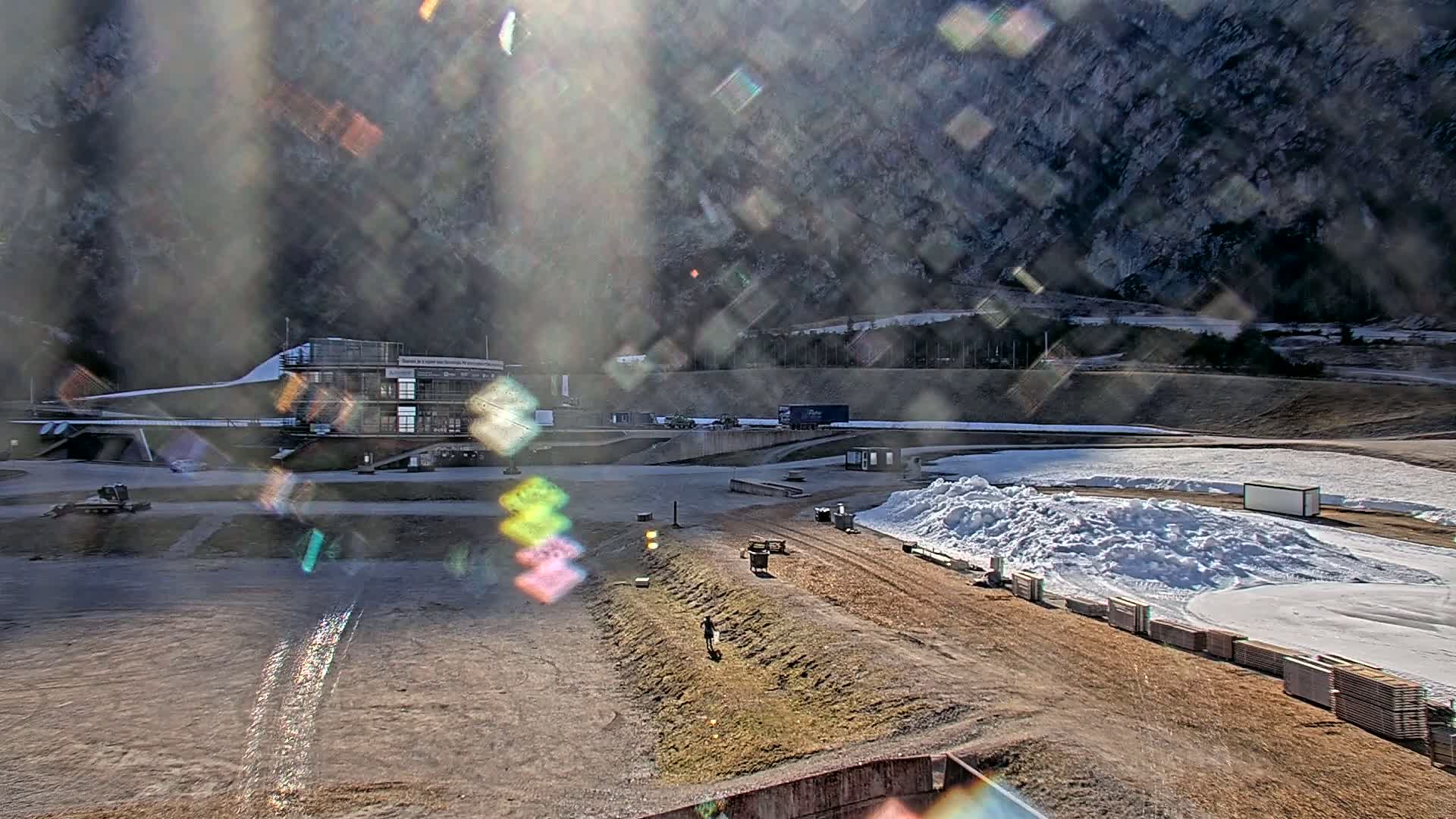 A sunny day reveals a snow-covered track with a building and equipment at a mountainside sports facility.