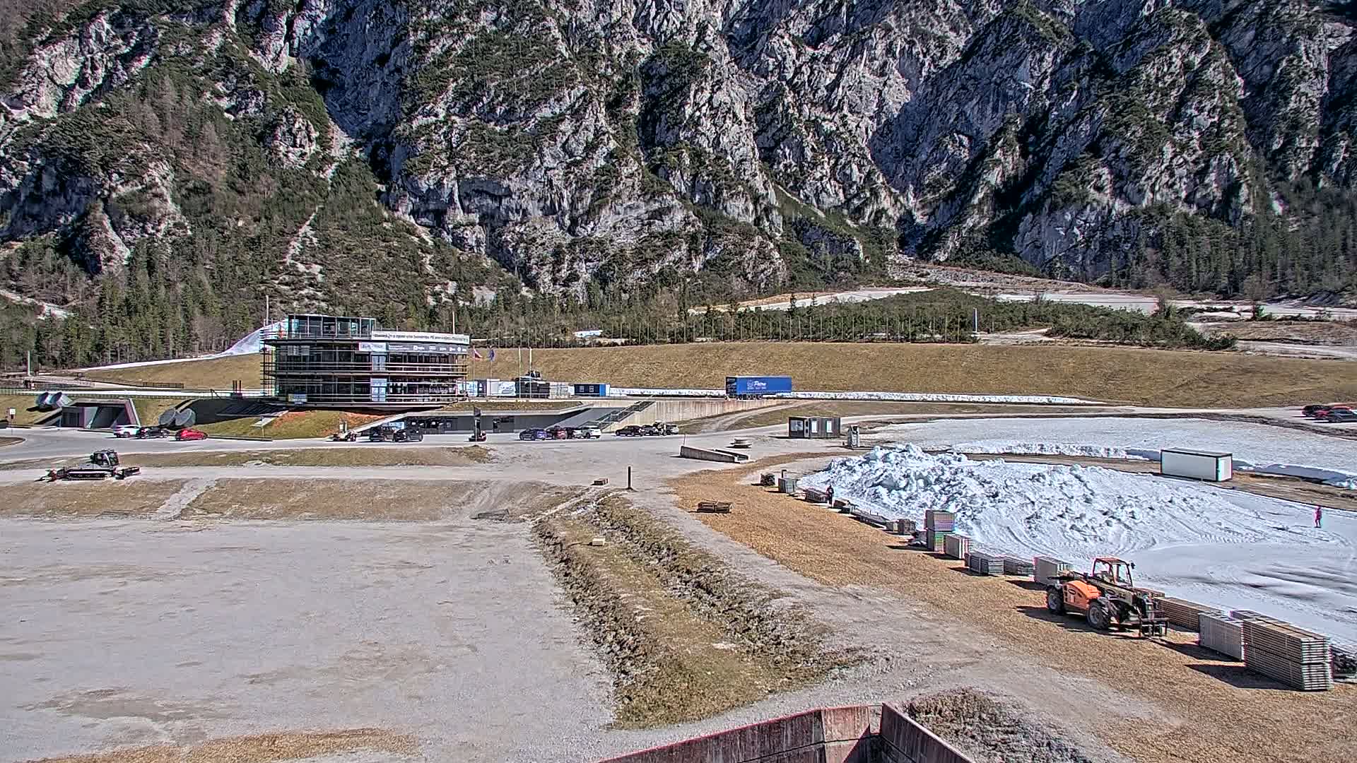A two-story building sits at the base of a large, rocky mountain on a sunny day, with a snow-covered area and several vehicles parked nearby.