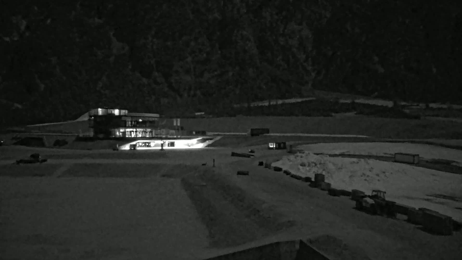 A brightly lit building sits at the base of a dark mountain at night, with snow and several pieces of machinery visible in the foreground.