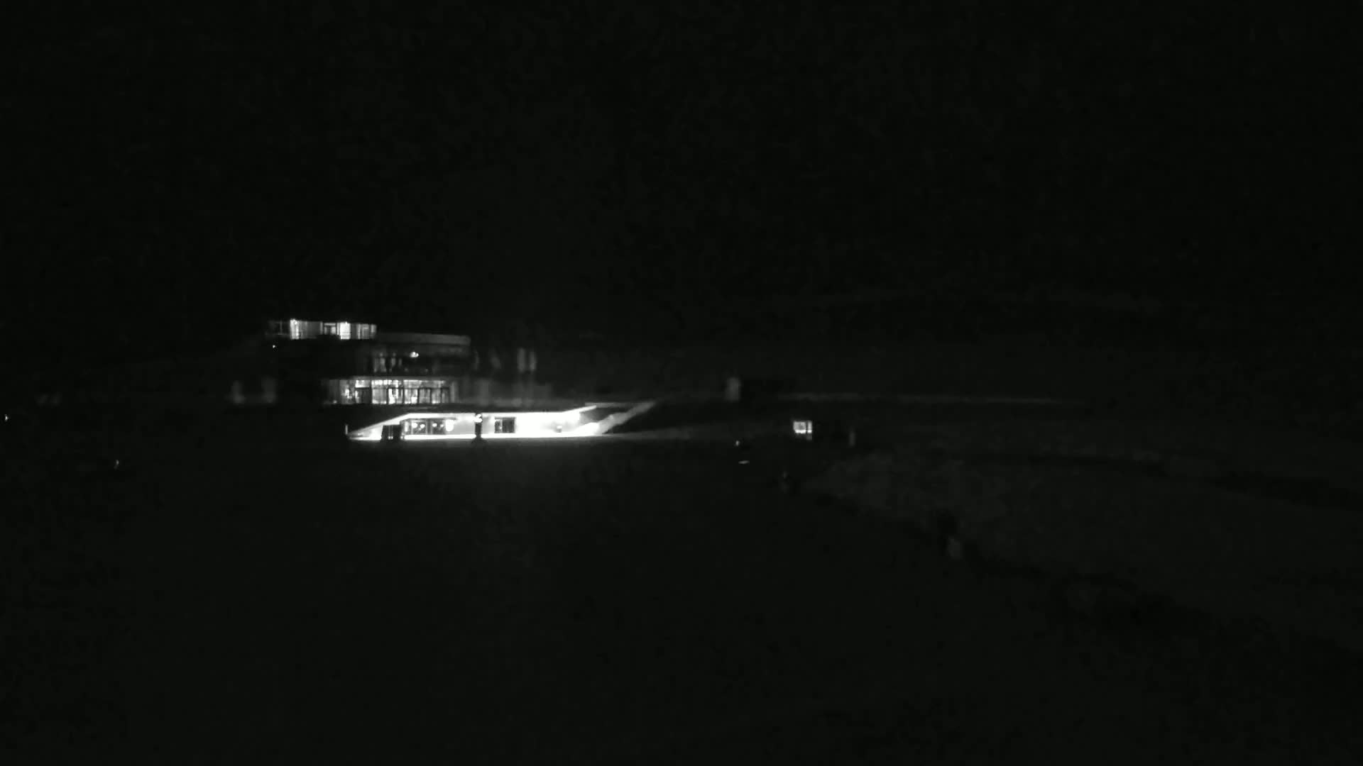 A brightly lit building is visible at night, with a dark foreground and faint lights suggesting vehicles or people in the distance.