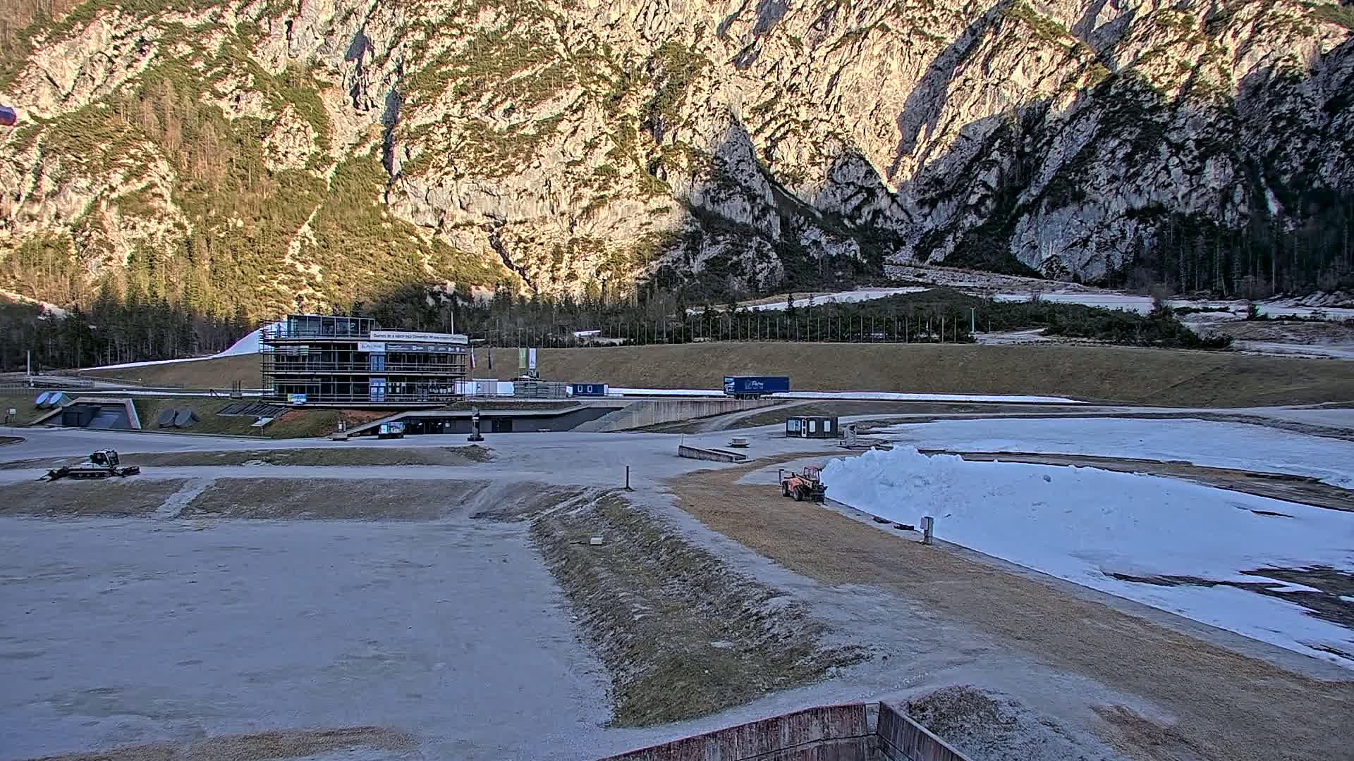 A bobsled track with a building and some machinery is situated in a valley before a large, rocky mountain under a mostly clear sky.