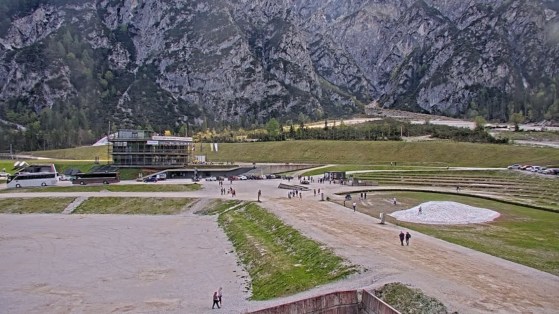 A large, open-air venue with a building and parking lot sits at the base of a mountain range on an overcast day.