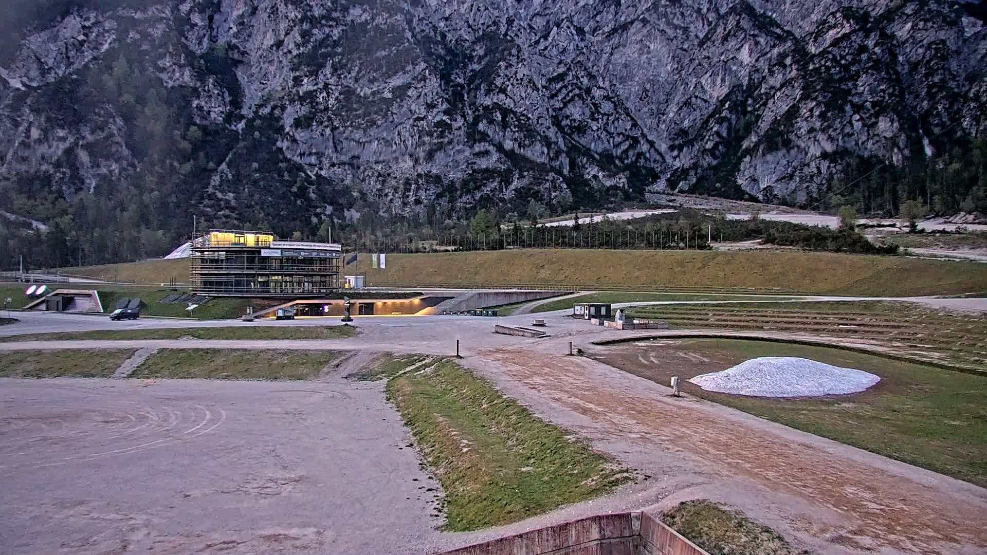 A modern building sits in a large, mostly empty outdoor area against the backdrop of a tall, grey mountain under a dusky sky.