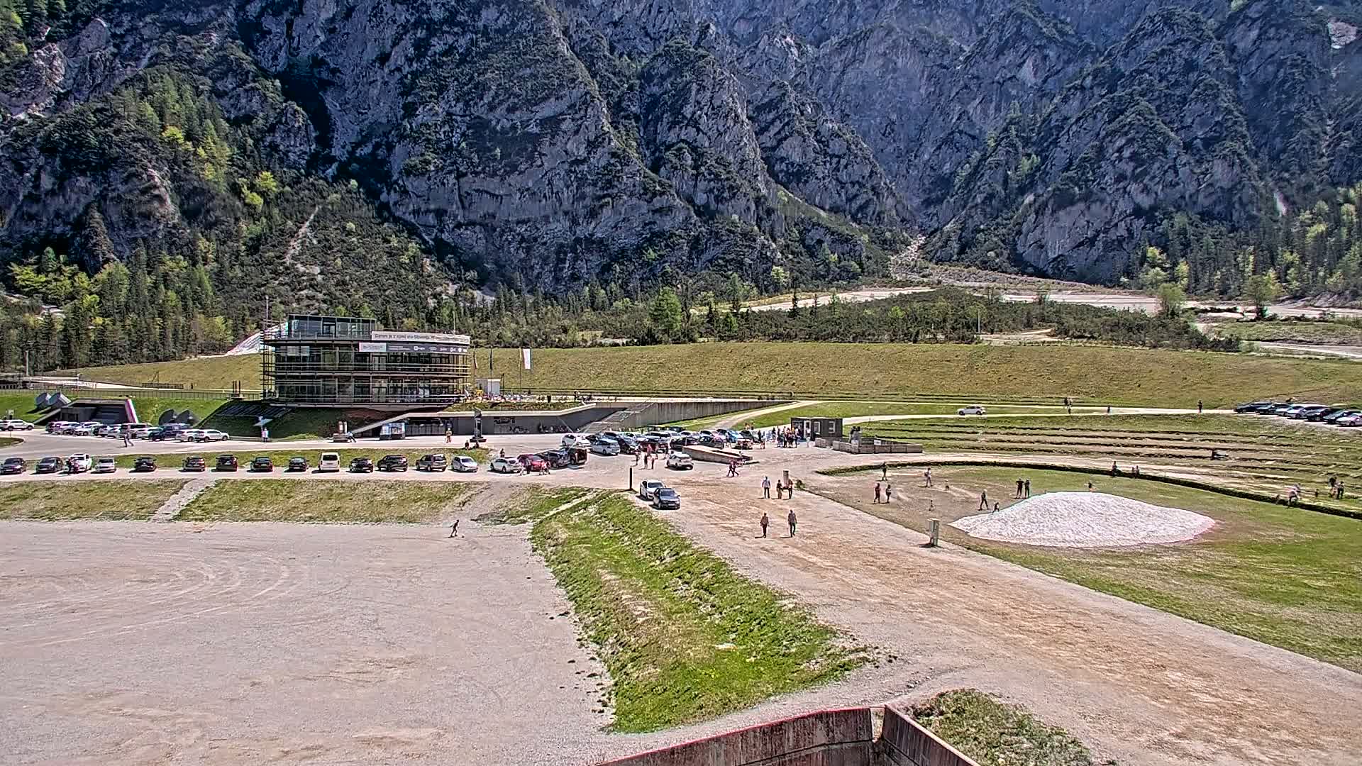 A parking lot filled with cars sits in front of a multi-story building at the base of a large, rocky mountain on a sunny day.