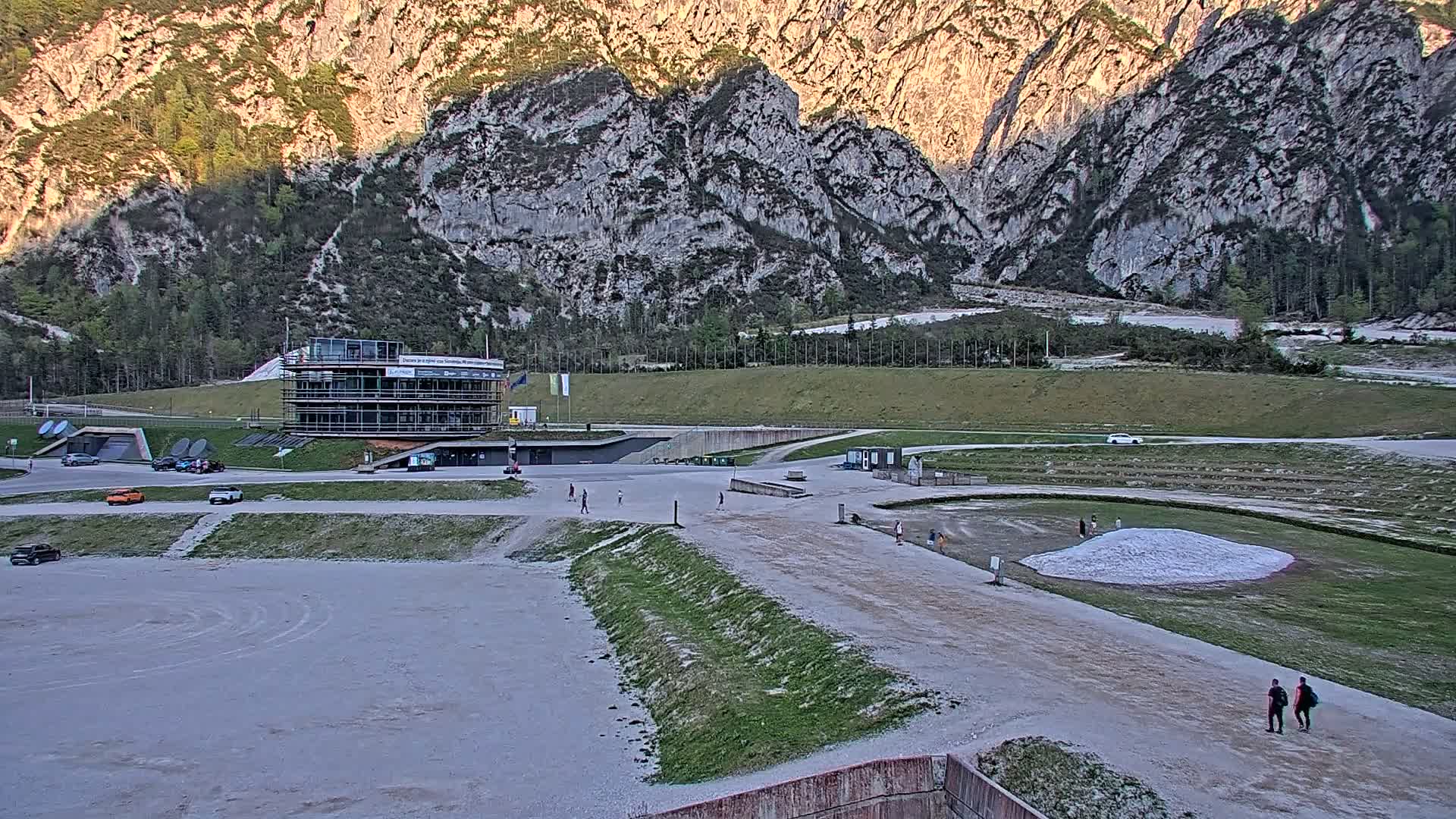 A large, light-grey, mostly empty paved area sits below a modern building and in front of a mountainous backdrop under a mostly clear sky.