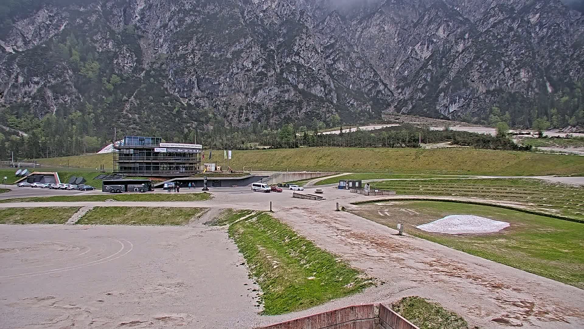 A large building sits near a mountainside in front of a wide, flat, partially grassy area with several vehicles parked nearby, under an overcast sky.