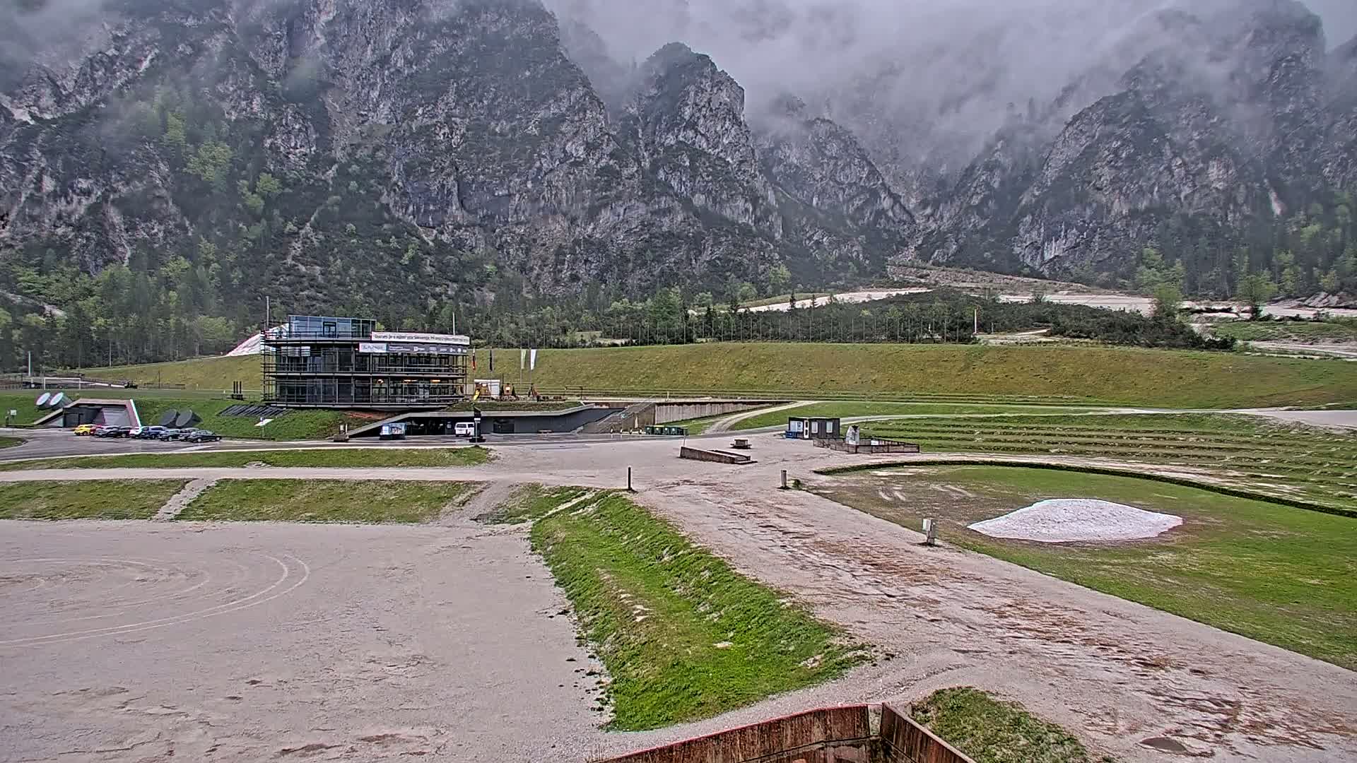 A modern building under construction sits in a valley surrounded by large, gray mountains under an overcast sky.